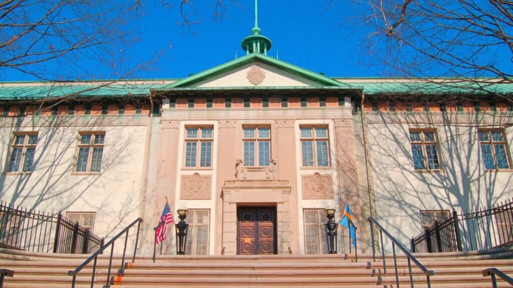 Front view of a historic building with a green roof, large wooden doors, and flagpoles on either side of the entrance, under a clear blue sky.