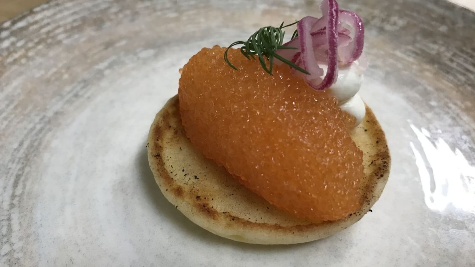 A small, round pancake topped with orange fish roe, a dollop of cream, thin red onion slices, and a sprig of dill on a textured plate.