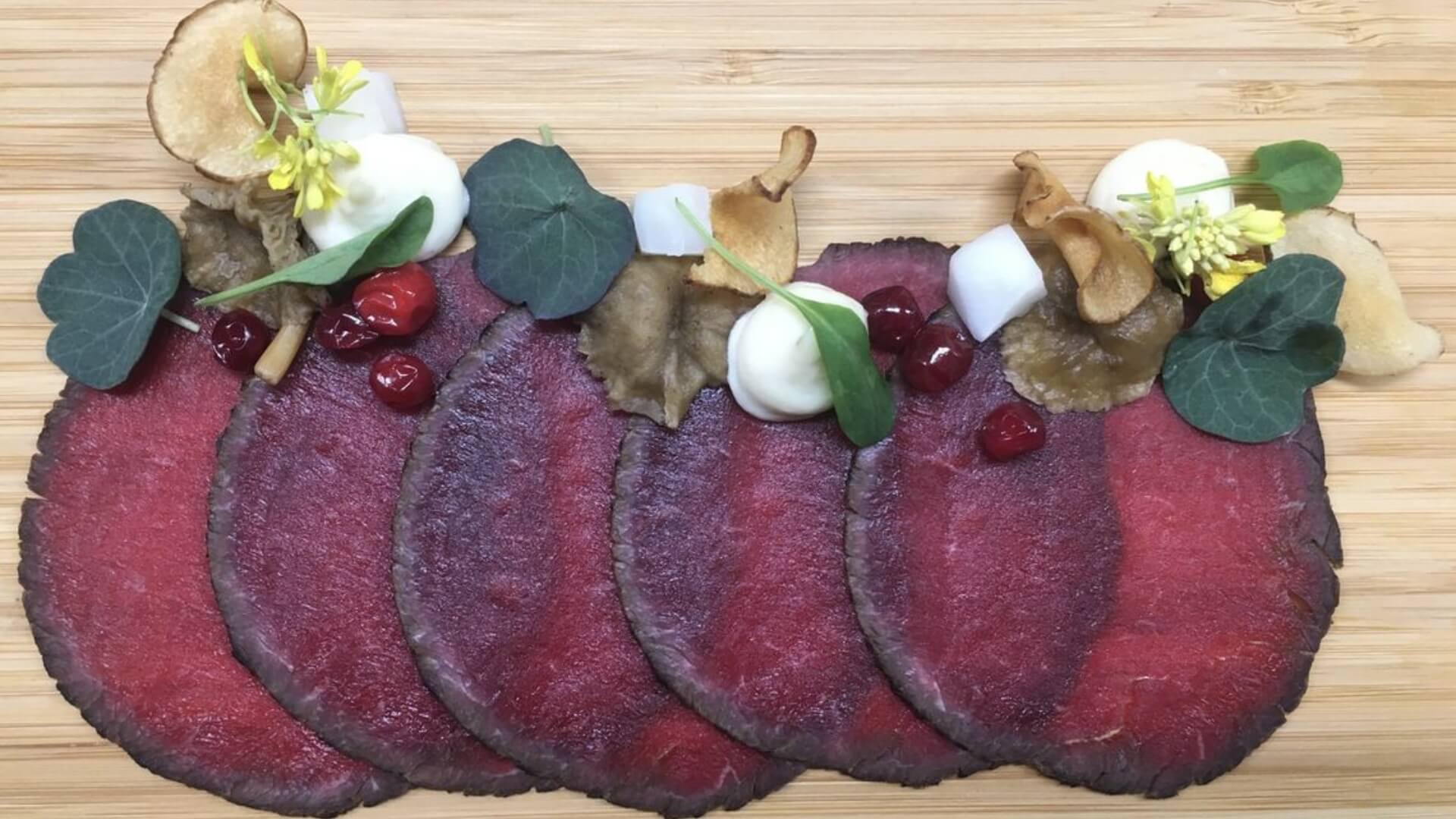 Slices of cured meat garnished with berries, dollops of cream, greens, yellow flowers, and crispy chips displayed on a wooden surface.