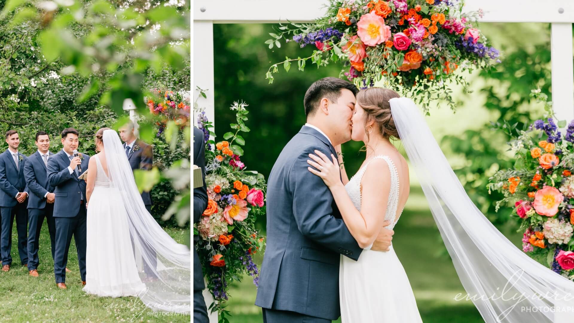 A couple kisses under a floral arch during their outdoor wedding ceremony. The bride wears a white dress with a long veil, and the groom is in a blue suit. Bridesmaids and groomsmen stand nearby.