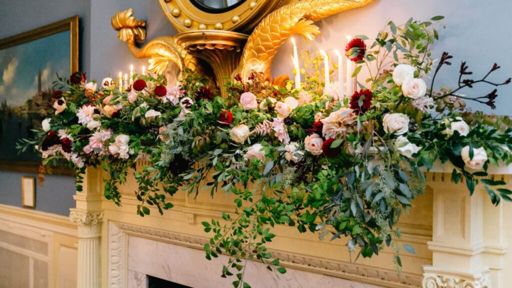 A fireplace mantel decorated with lush greenery, pink and red flowers, candles, and a golden decorative piece above it.