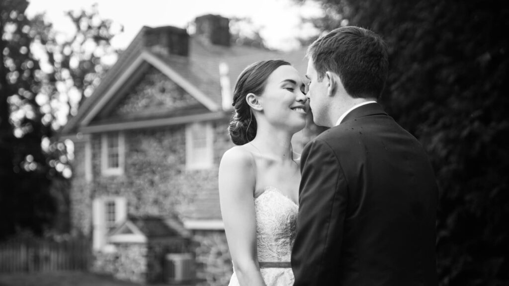 A bride and groom share a moment outside, standing close together, with a stone house in the background.