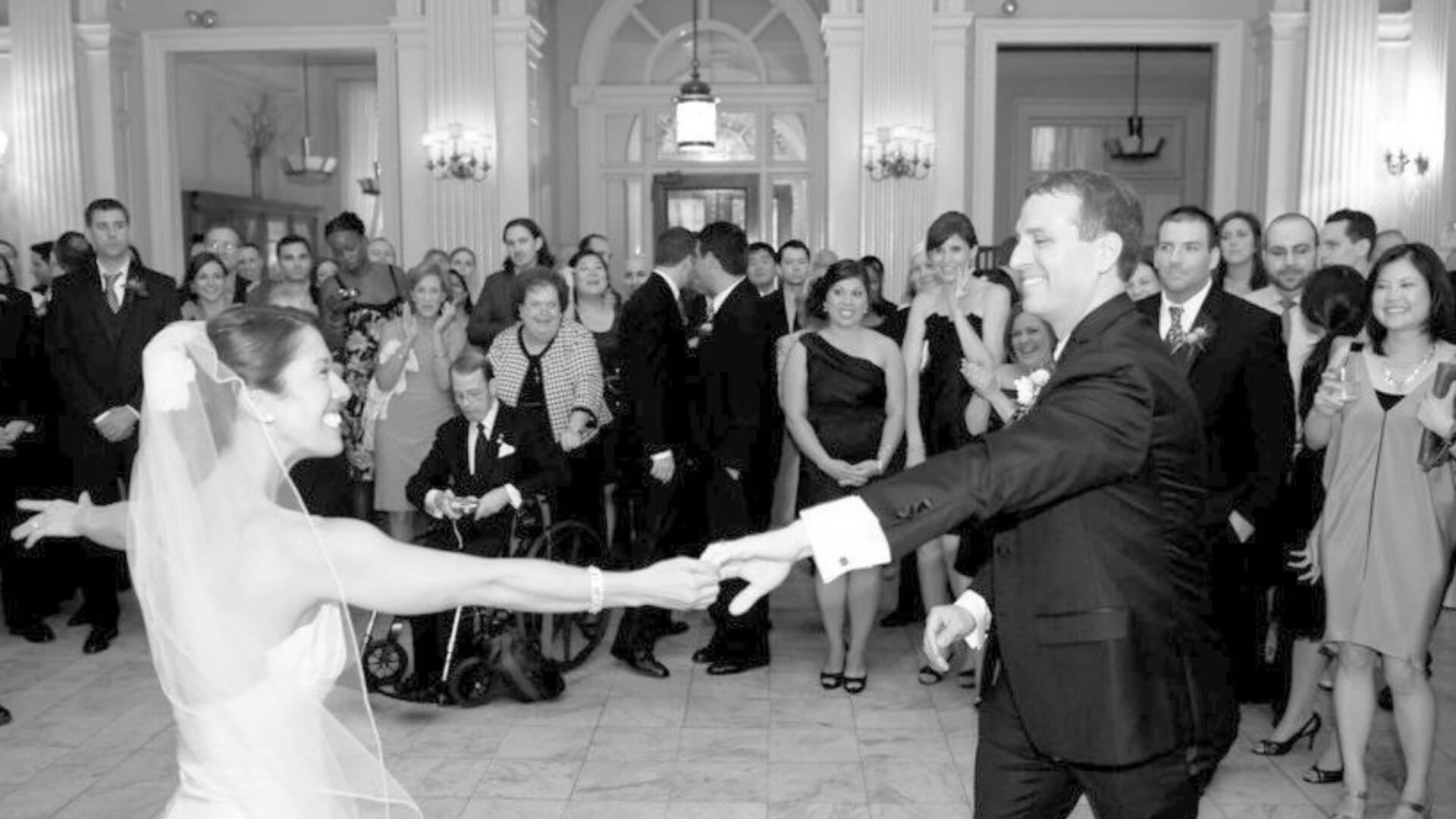 A bride and groom dance in a formal setting, surrounded by seated and standing guests watching them.