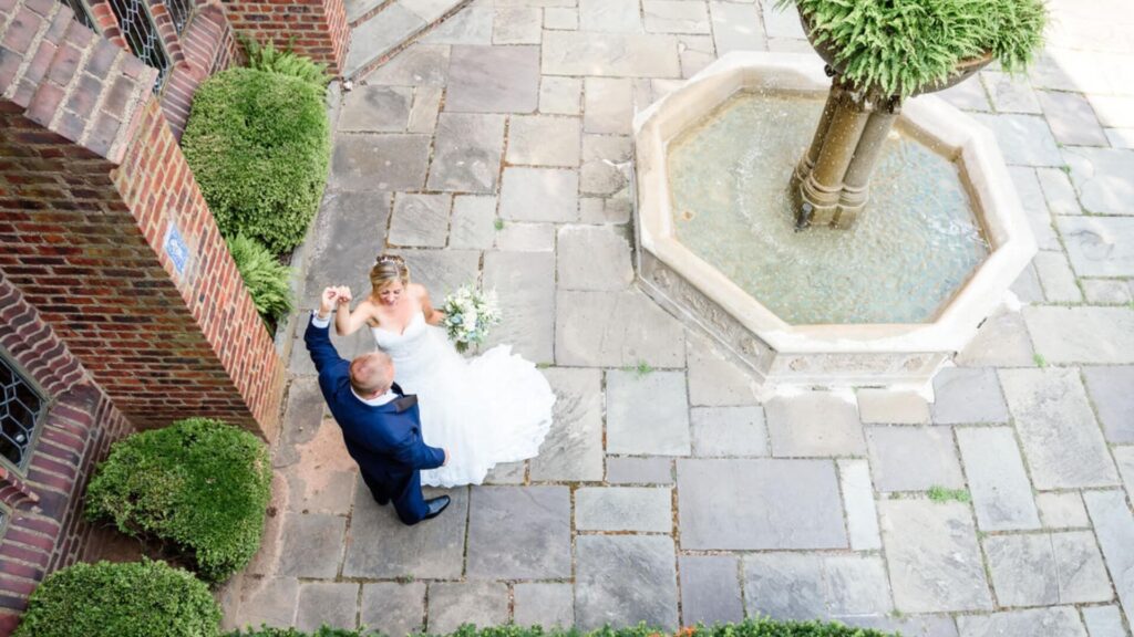 Bride and groom dance on a stone patio beside a fountain, enclosed by brick walls and greenery.