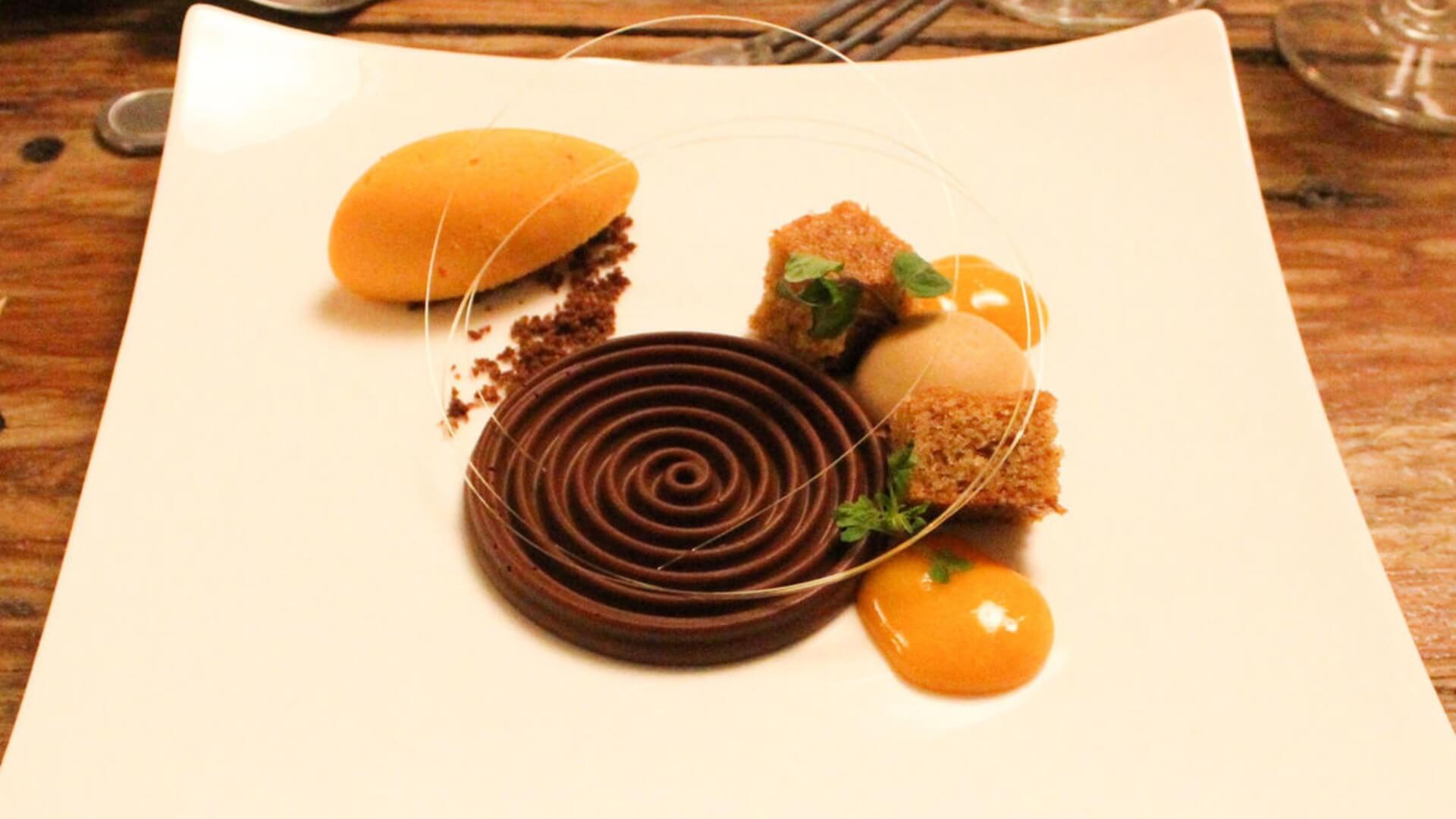 A gourmet dessert featuring a chocolate spiral, orange sorbet, sponge cake cubes, and garnished with herbs on a white plate.