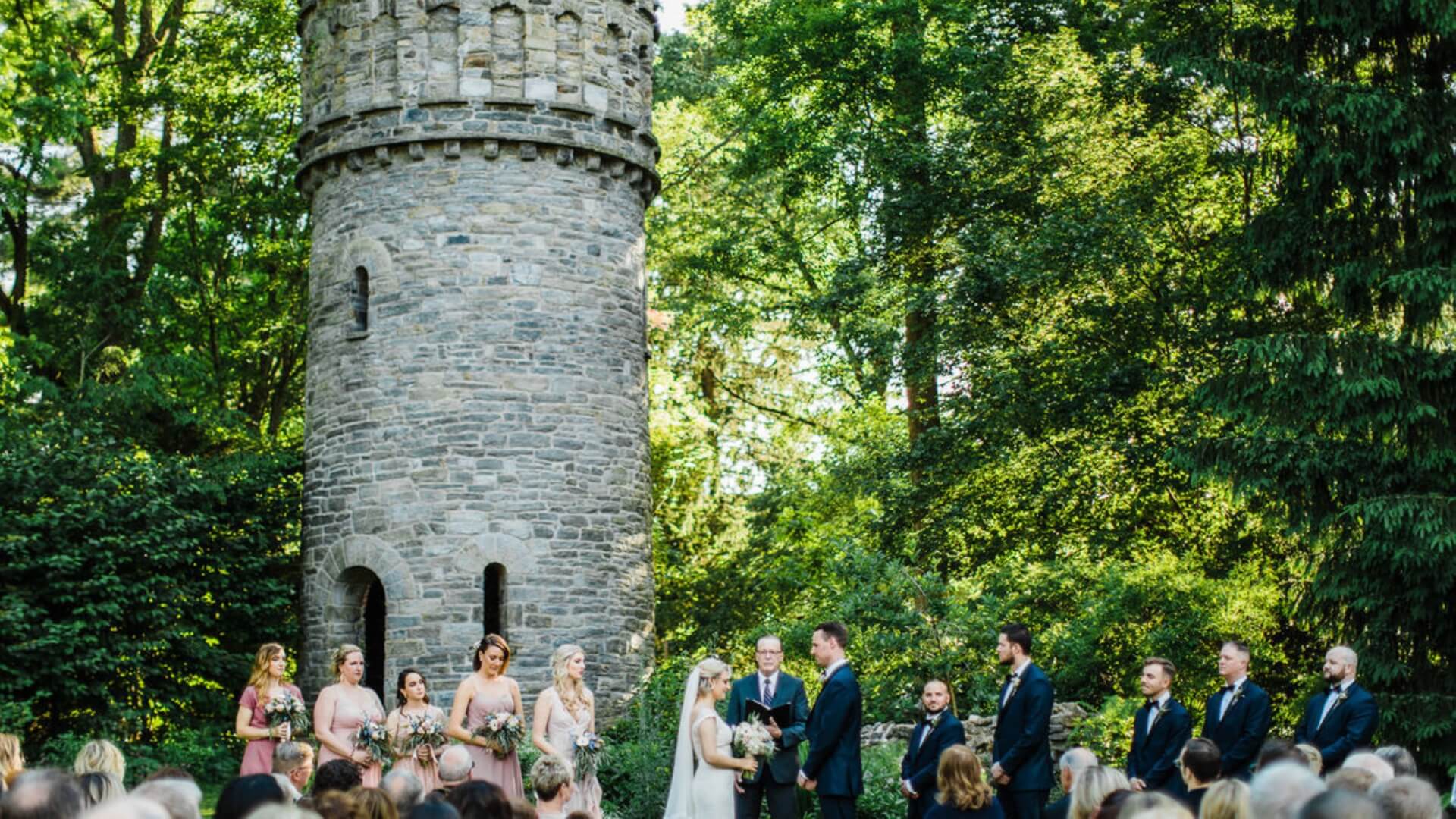 Outdoor wedding ceremony with a couple exchanging vows in front of a stone tower, surrounded by bridesmaids and groomsmen, with guests seated in the foreground.