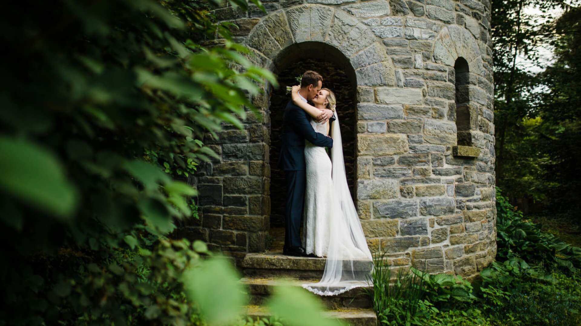 A couple embraces on the stone steps of a round stone structure surrounded by lush greenery.