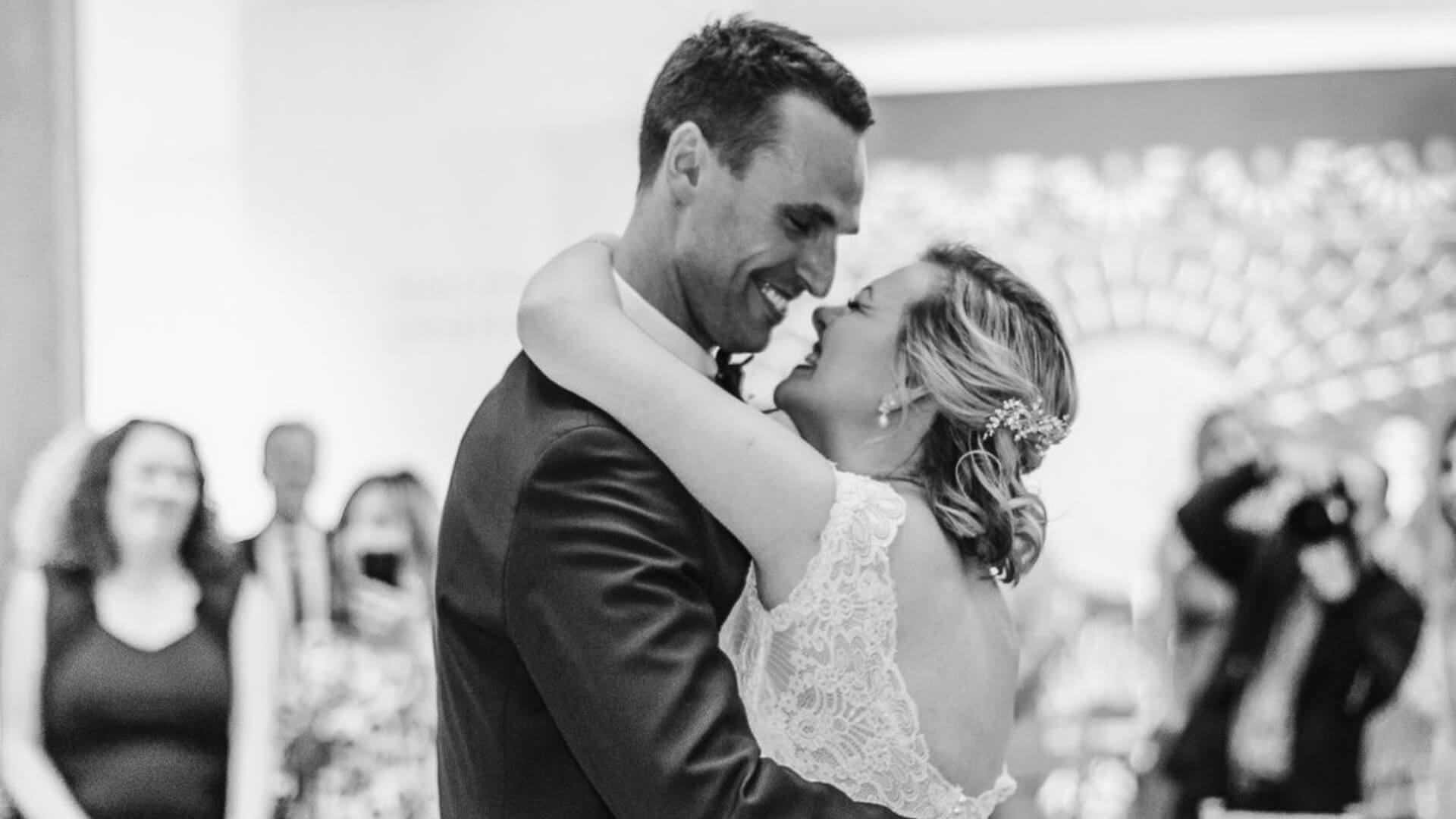 A bride and groom embrace and smile at each other during their wedding dance, with guests watching in the background.