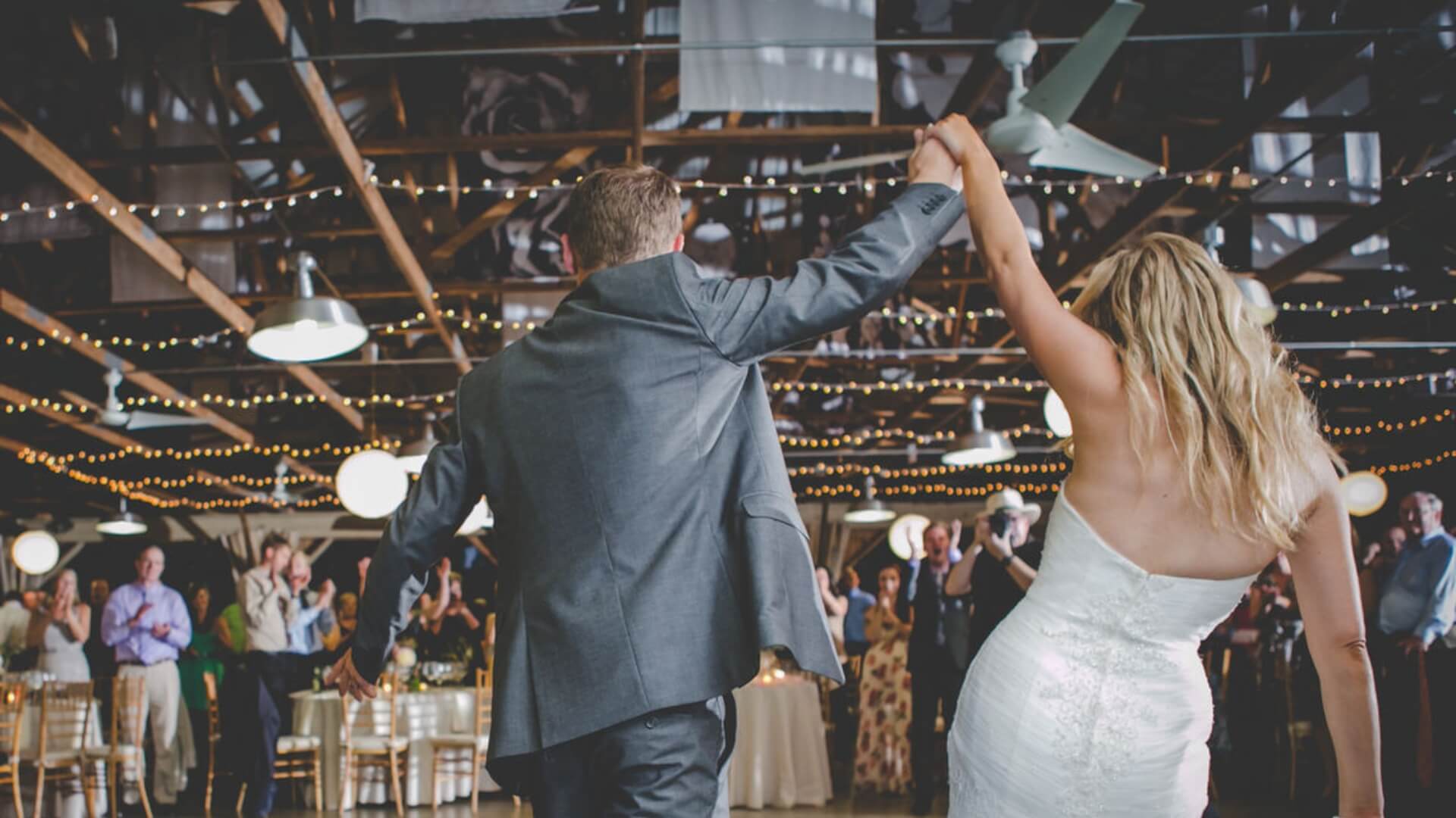 A couple in wedding attire enters a decorated reception hall, holding hands with arms raised. Guests stand and applaud.