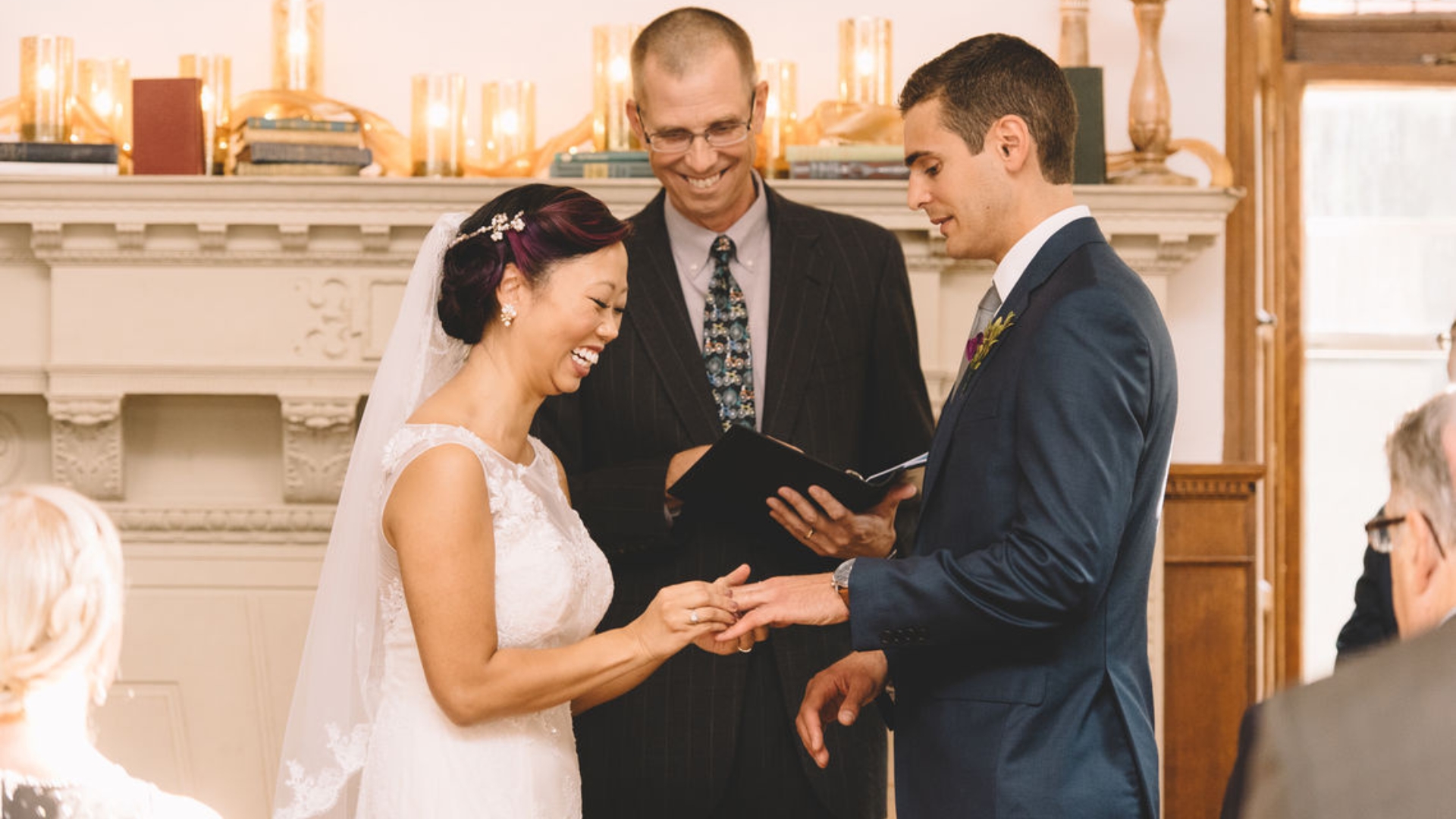 A bride and groom exchange rings during a wedding ceremony, with an officiant smiling behind them. Guests are seated nearby, and candles are displayed on a mantel in the background.