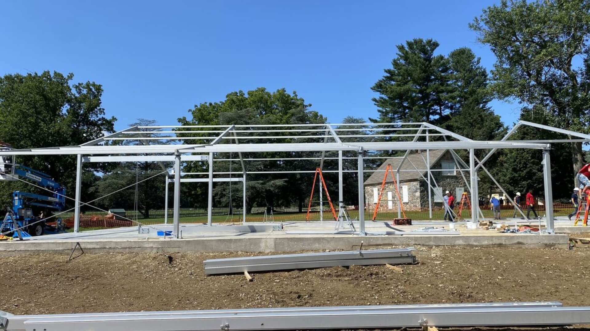 Construction site with a metal framework for a building, surrounded by trees and a house in the background, under clear blue sky. Workers and tools are visible.