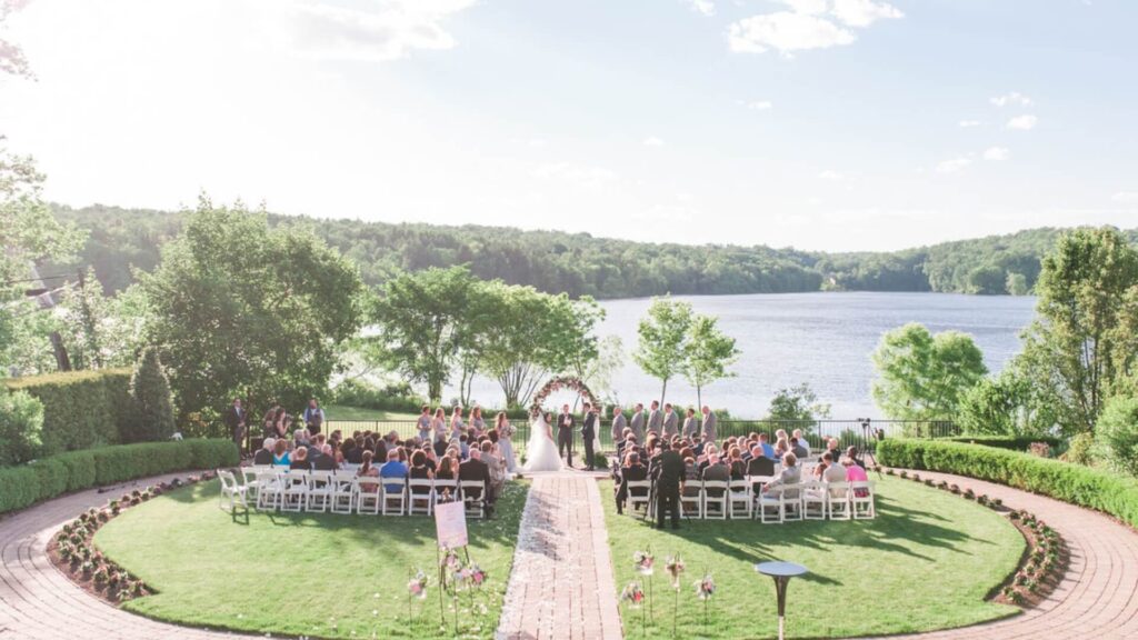 Outdoor wedding ceremony by a lake, featuring guests seated on white chairs, a flower arch, and a lush green landscape under a clear sky.