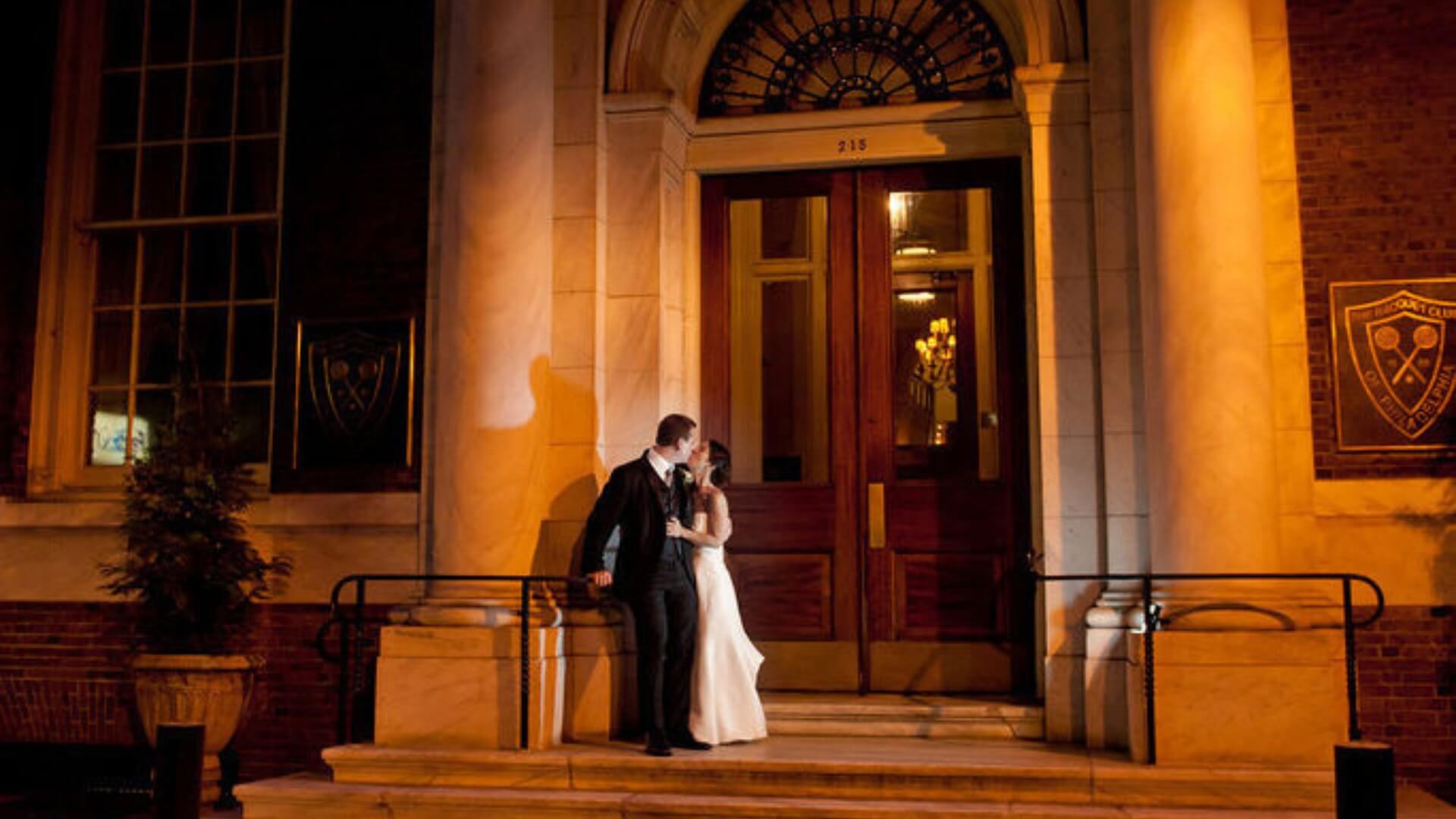 A couple in formal attire stands on the steps of a large building at night. The lighting casts long shadows, and the entrance features large wooden doors and decorative arches.