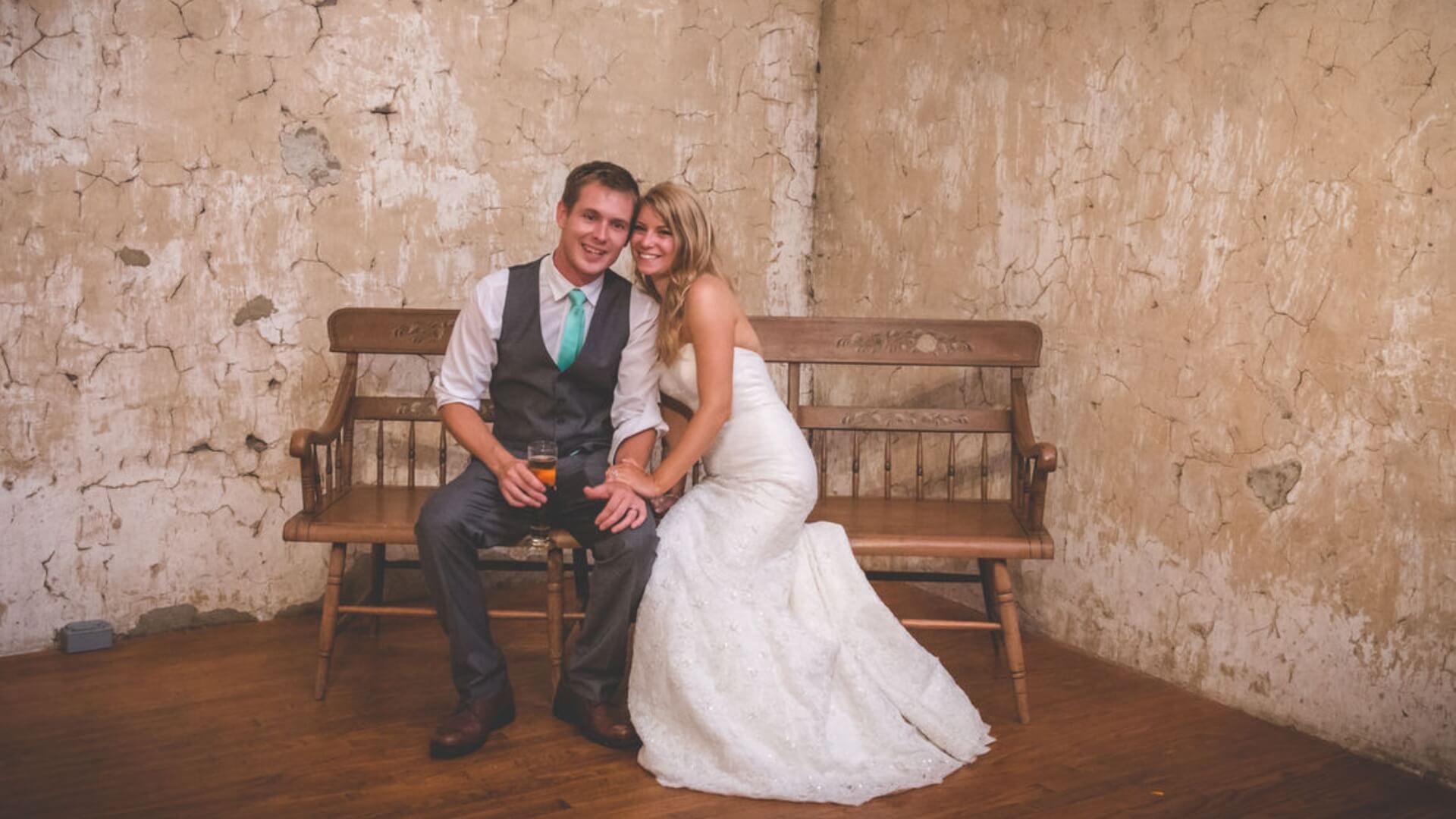 A bride in a white gown and a groom in a vest and tie sit on a wooden bench against a weathered, cracked wall, smiling and holding a drink.