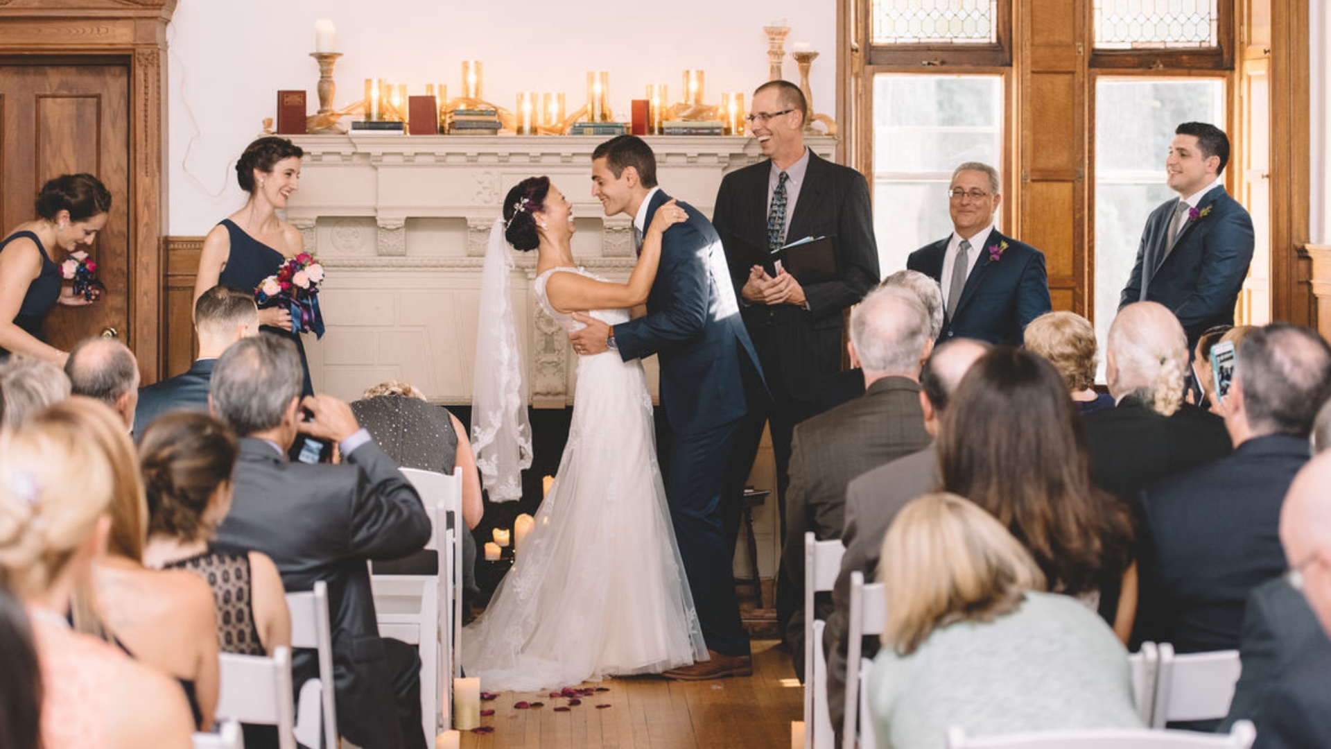 Couple exchanges vows at a wedding ceremony. The bride and groom stand at the altar, surrounded by the officiant, bridal party, and guests seated in rows.