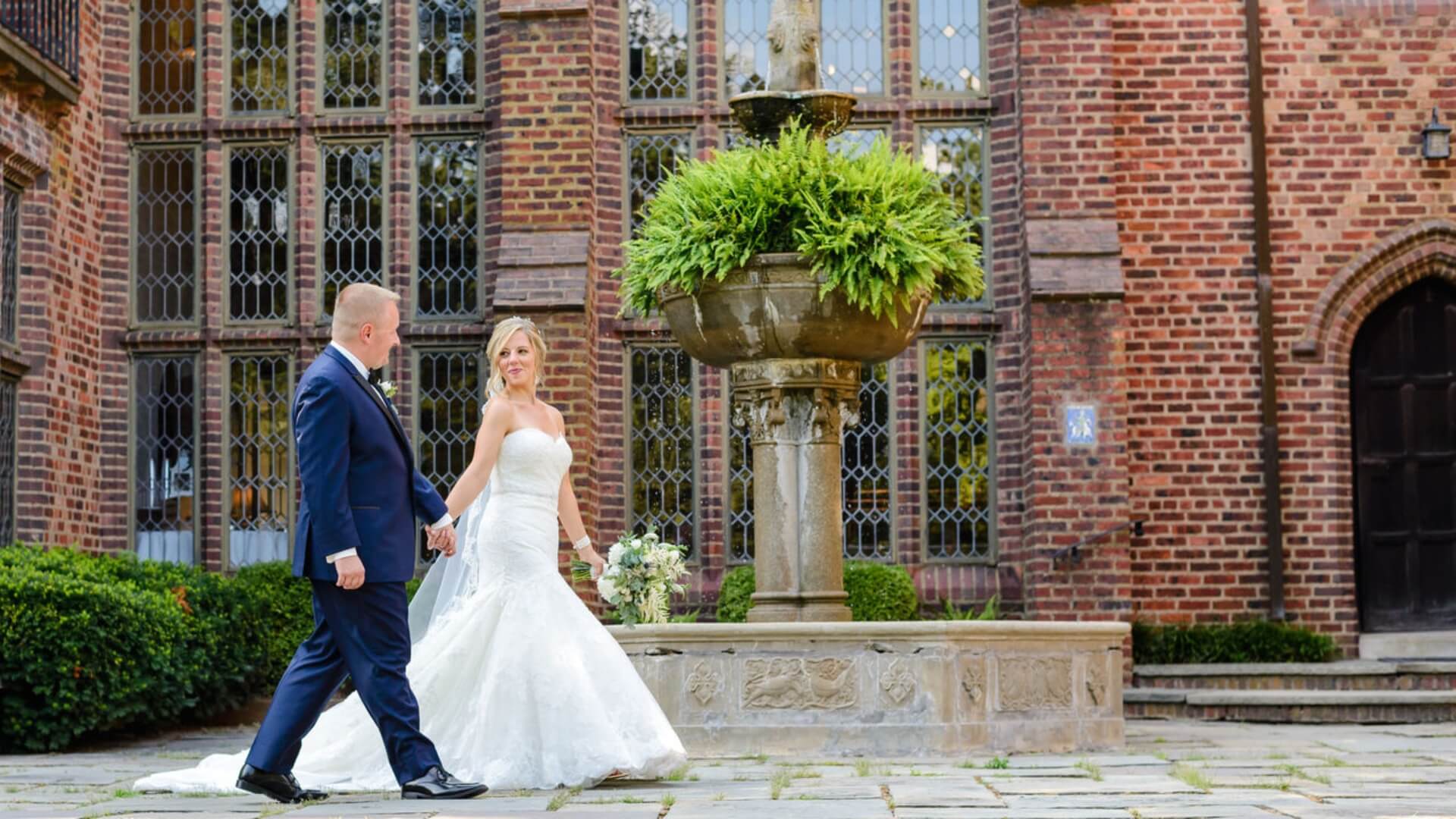 A bride and groom walk hand-in-hand near a stone fountain and a brick building with large windows.