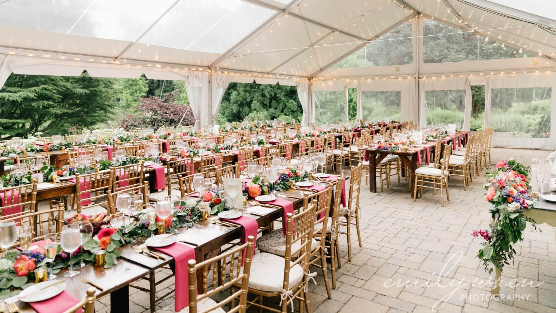 Outdoor wedding reception under a transparent tent with wooden chairs and tables. Tables are adorned with pink napkins, floral centerpieces, and string lights overhead.