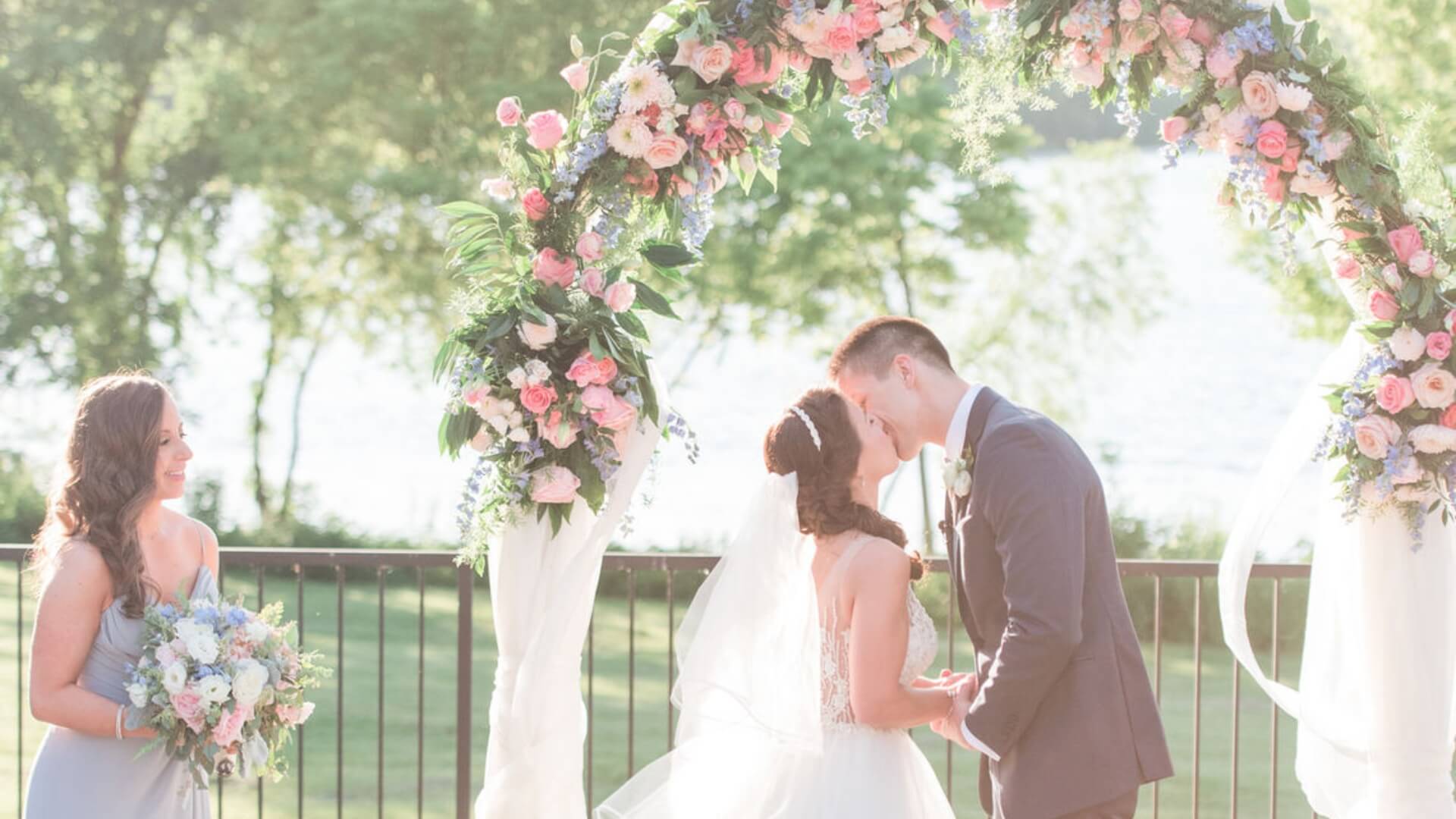 A couple kisses at an outdoor wedding under a floral arch, with a bridesmaid holding a bouquet nearby.