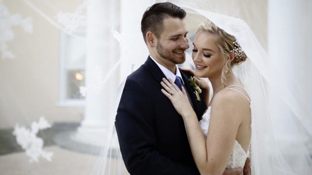 A bride and groom embrace, both smiling, in wedding attire under a sheer veil.