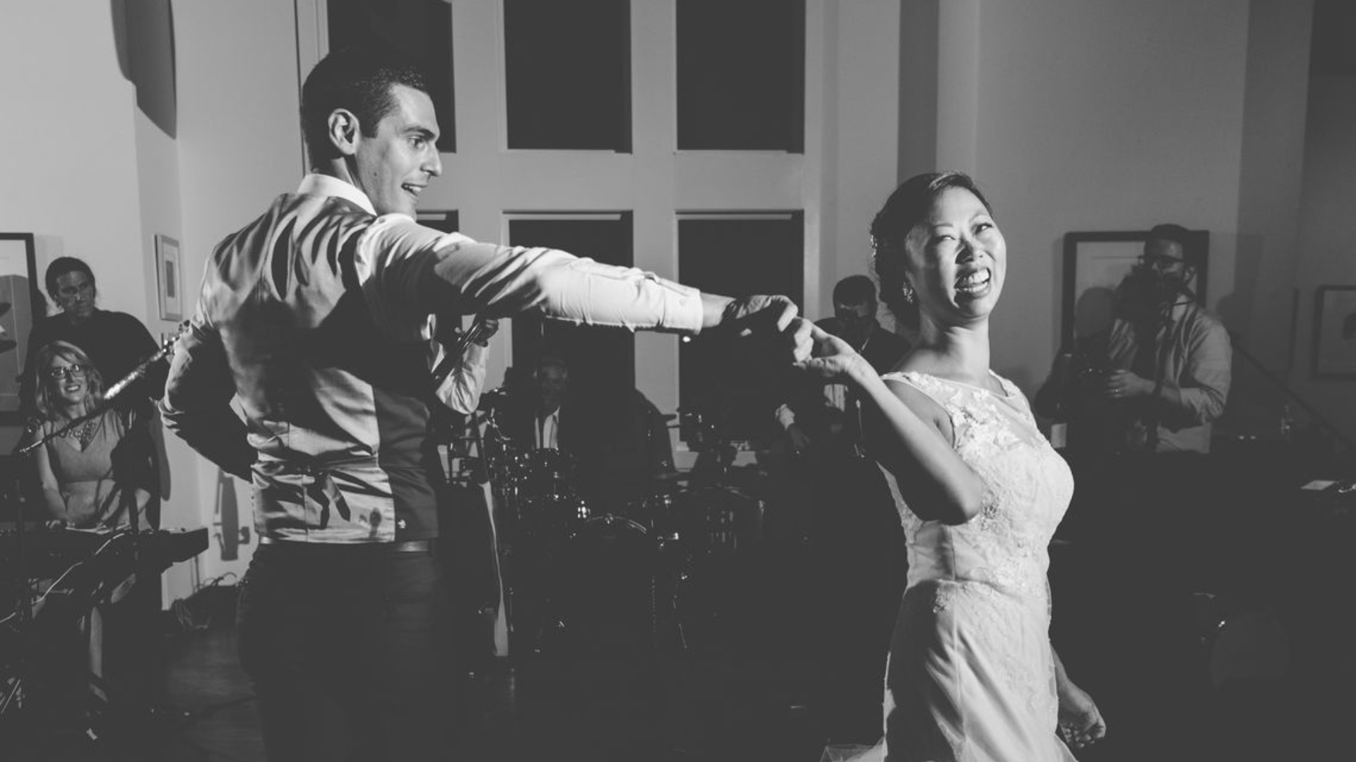 A couple dances joyfully in a room with large windows and band equipment in the background. The photo is in black and white.