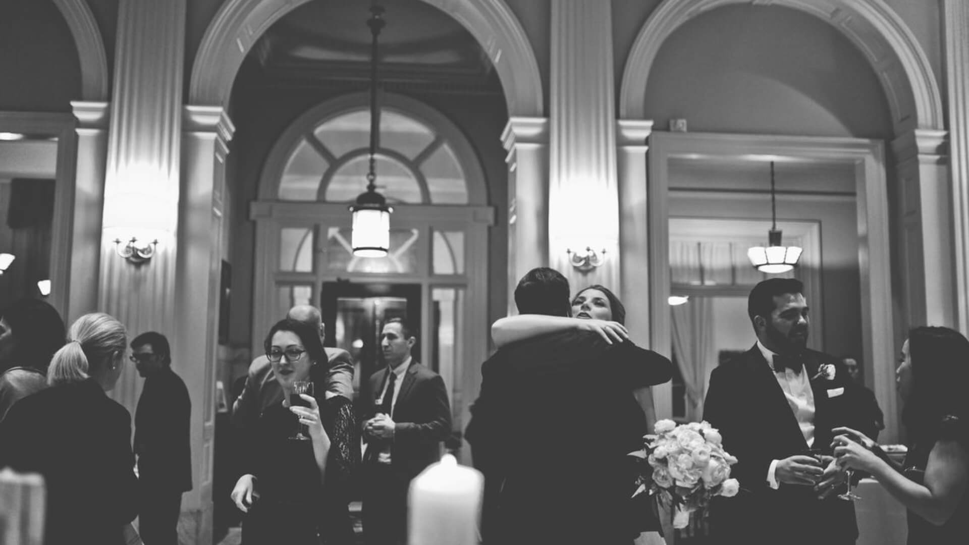 People mingle and embrace in a formal setting with tall arches and columns. The scene is black and white, with guests holding drinks and flowers.