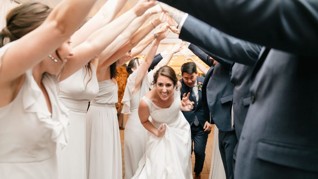 Bride and groom smiling as they walk through an arch of raised arms formed by guests in formal attire.