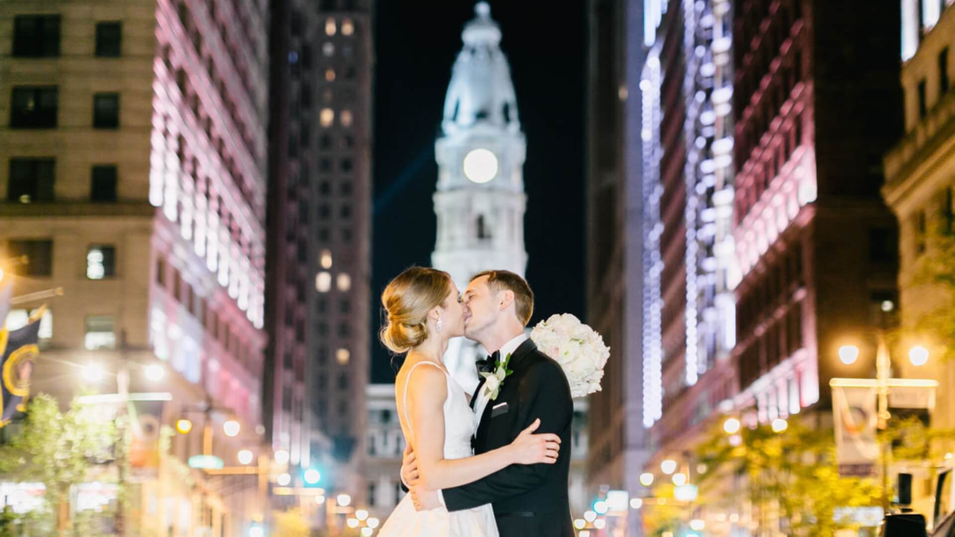 A bride and groom kiss on a city street at night, with a tall, illuminated building in the background.