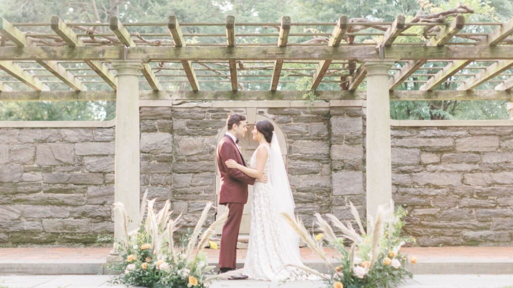 A couple in wedding attire stands facing each other under a wooden pergola, surrounded by greenery and floral arrangements.
