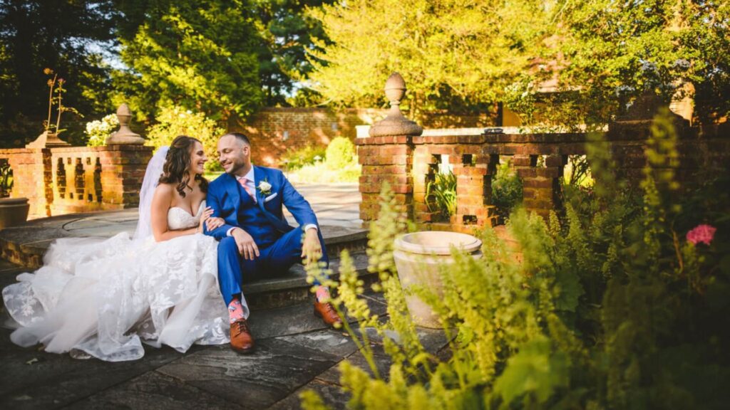 A bride in a white dress and a groom in a blue suit sit on stone steps, surrounded by greenery and brick architecture, smiling at each other.
