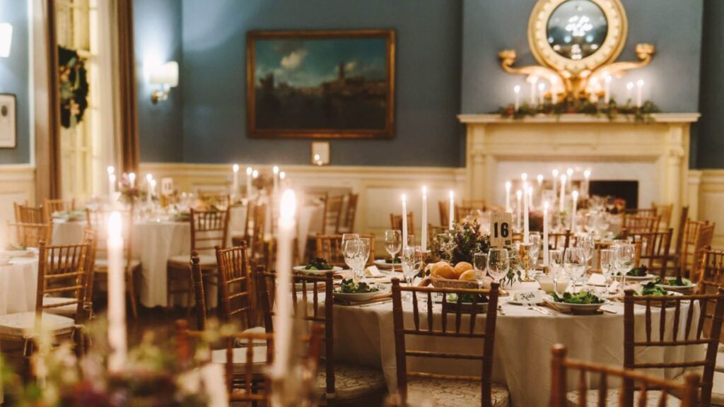 Elegant dining room set for an event with round tables, white tablecloths, candles, and wooden chairs. A fireplace and paintings adorn the blue walls in the background.