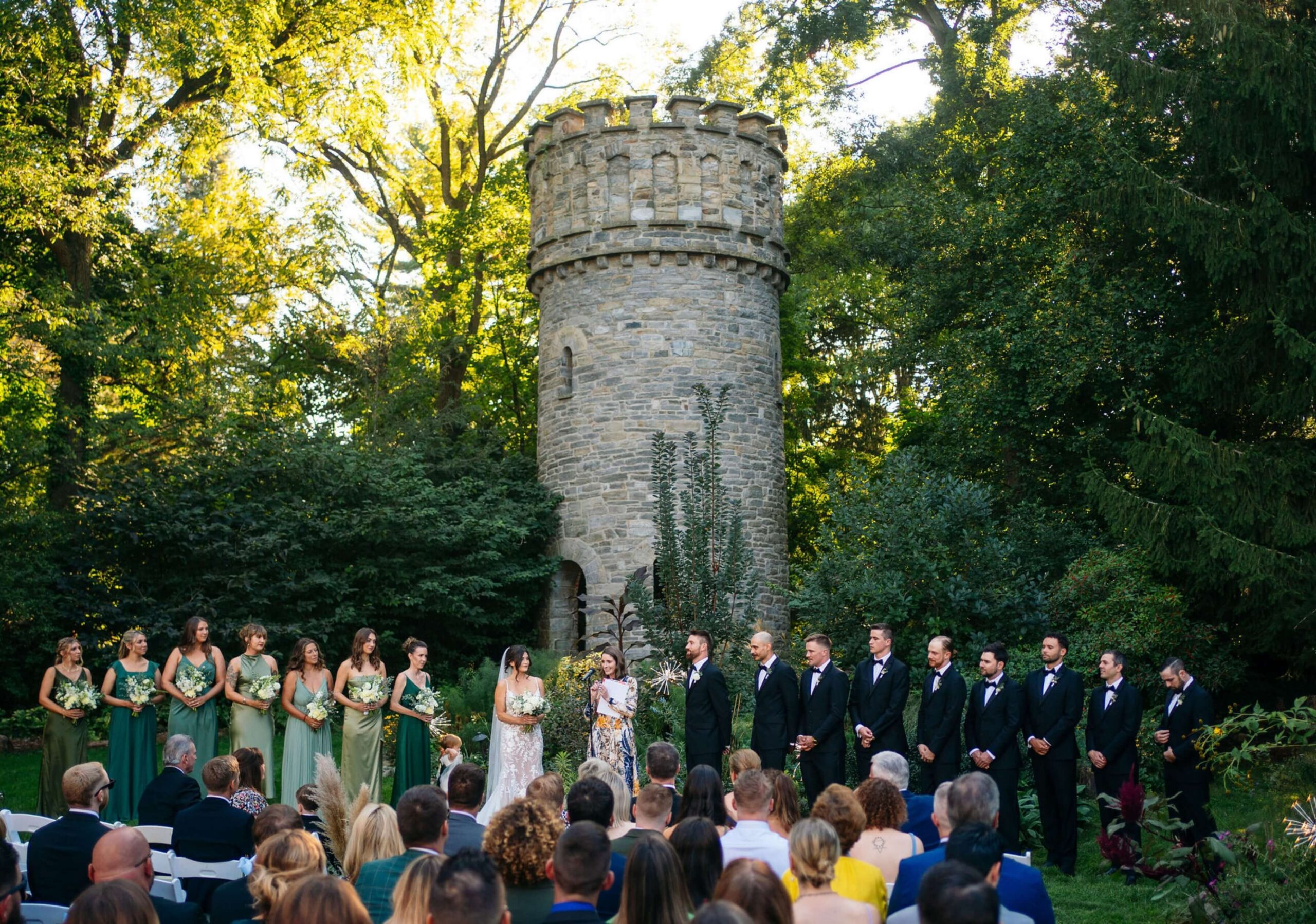 A wedding party stands in front of a stone tower surrounded by greenery. Bridesmaids wear green dresses, and groomsmen wear dark suits. Guests are seated under a canopy of trees.