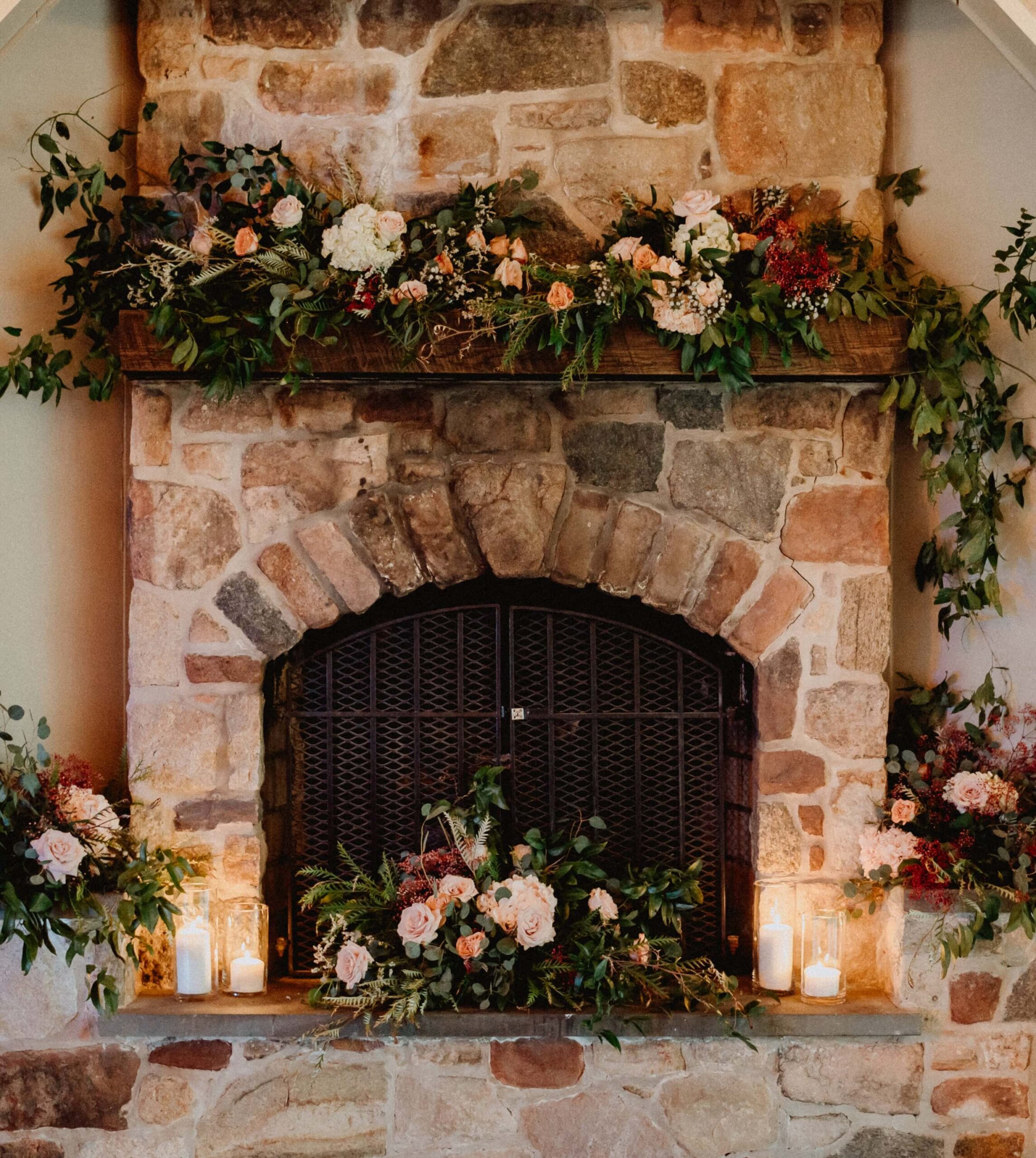 A stone fireplace decorated with greenery, pink and white flowers, and lit candles.
