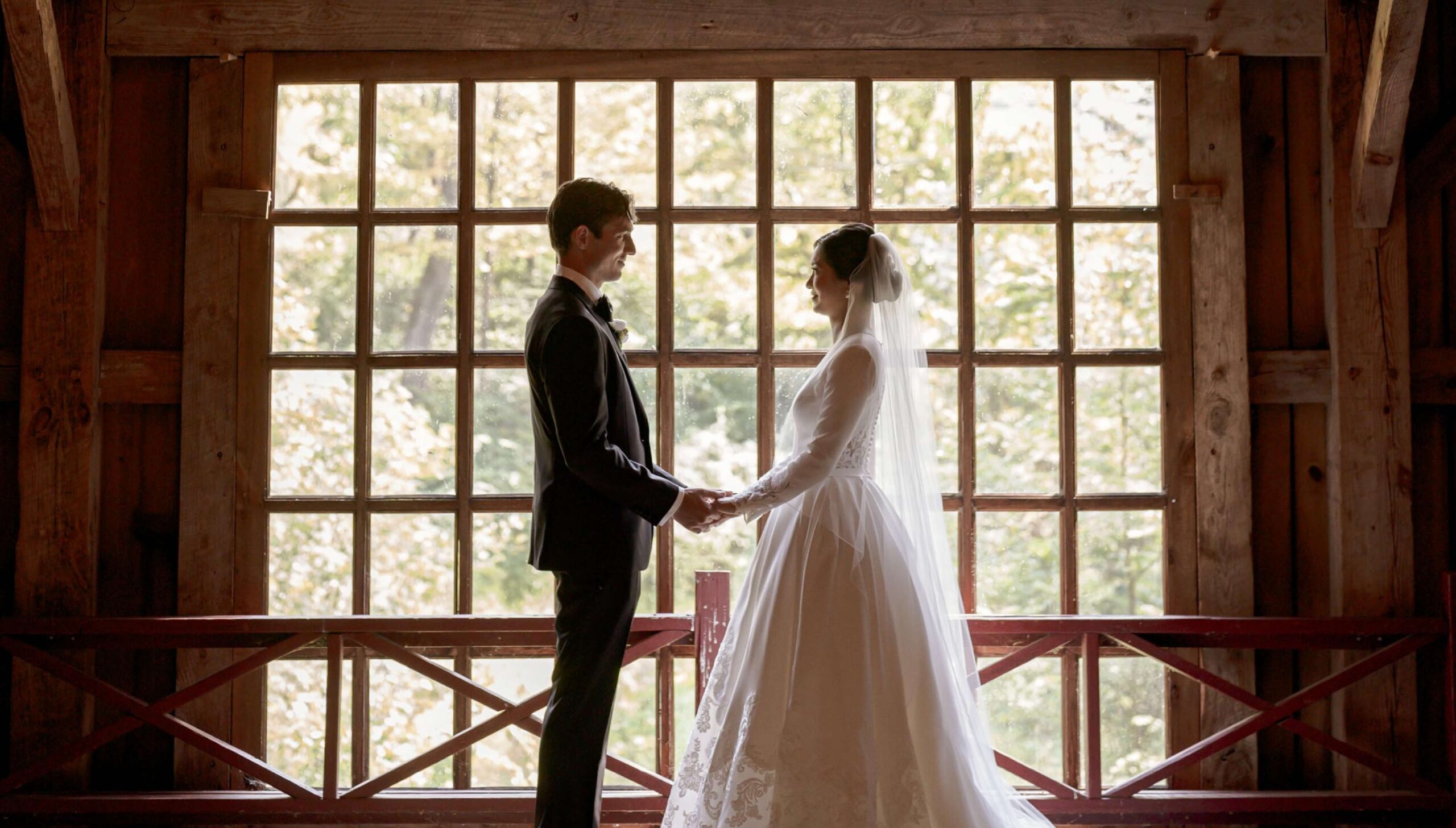 A bride and groom stand facing each other, holding hands, in front of large windows with natural light streaming through.