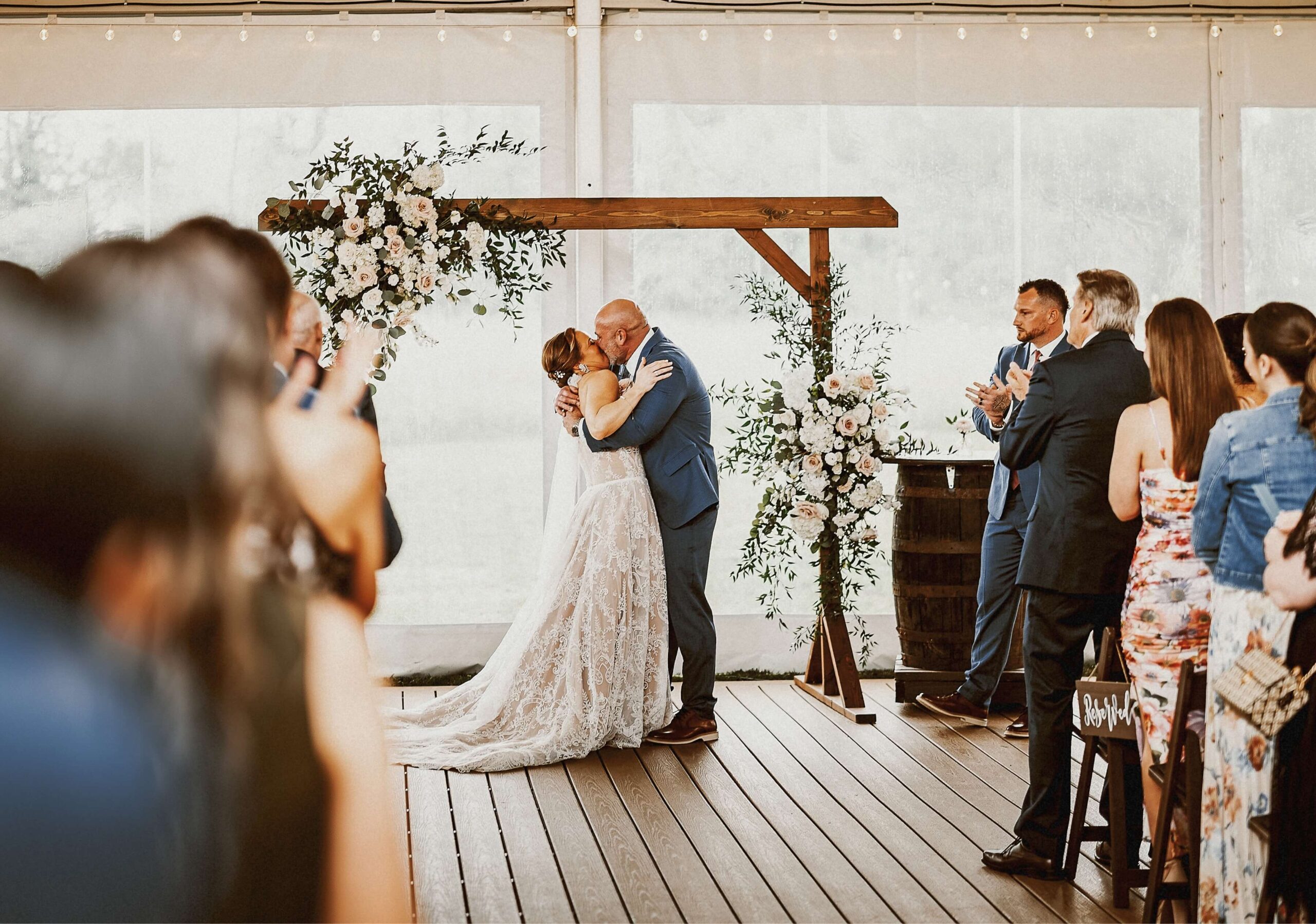 A couple embraces under a floral arch at a wedding ceremony, surrounded by guests.