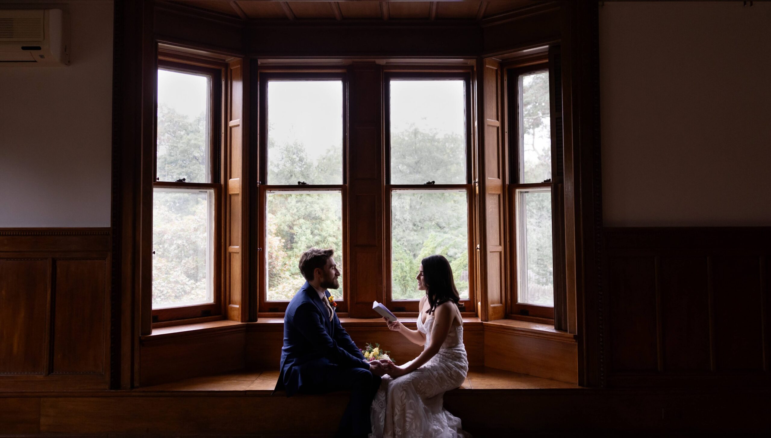 A couple in formal attire sits on a wooden window seat in a room with three large windows. They face each other with greenery visible outside.