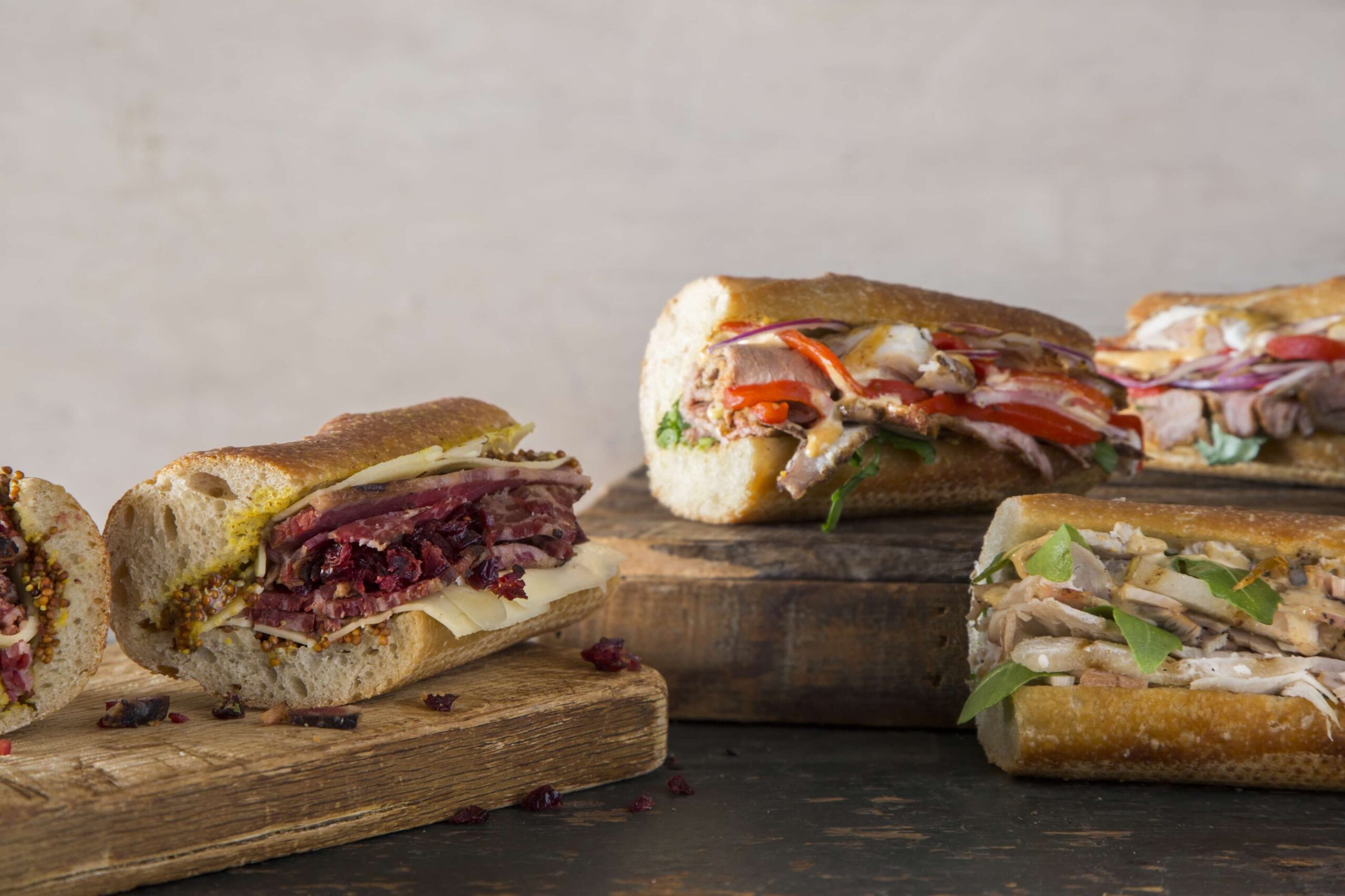 Three sandwiches on rustic wooden boards, each filled with various meats, cheeses, and vegetables, displayed against a neutral background.