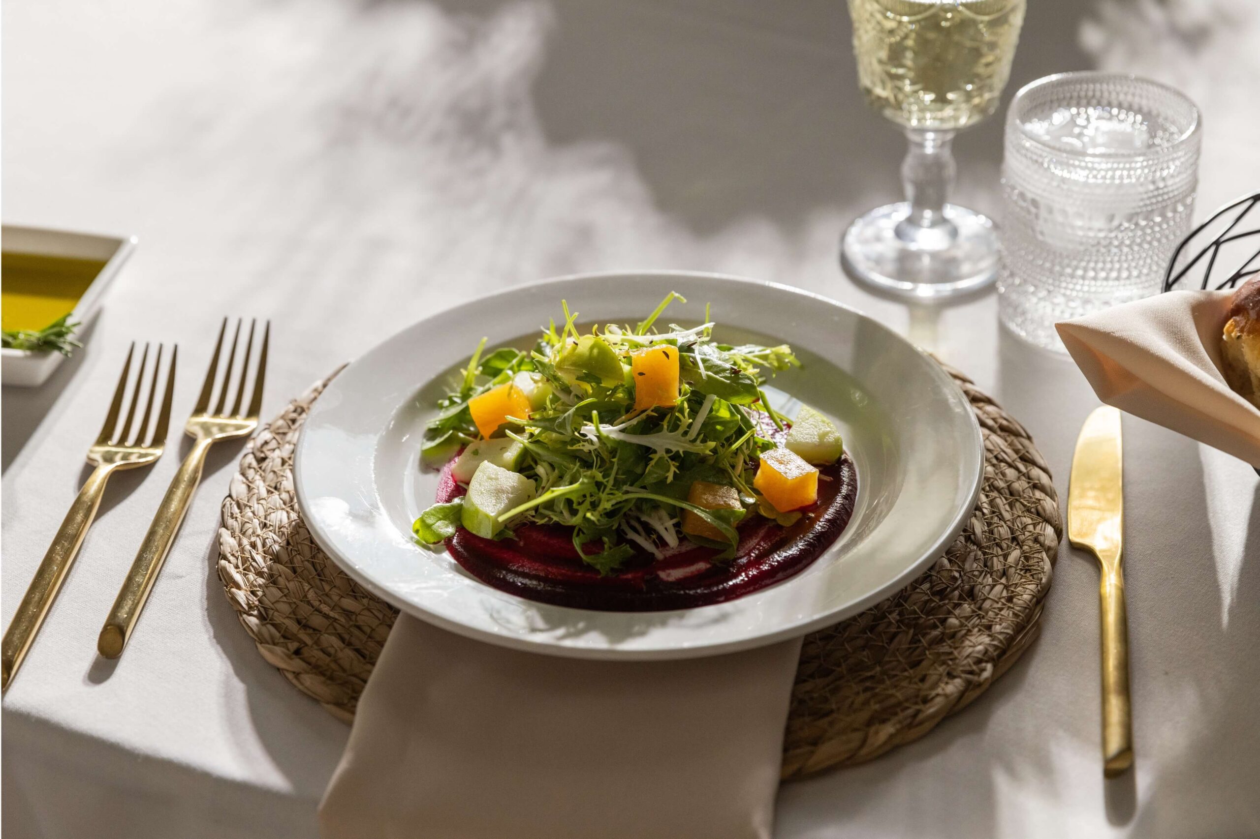 A plated salad with arugula, diced yellow and white vegetables, and beet slices sits on a woven placemat, surrounded by gold cutlery and a glass of white wine.
