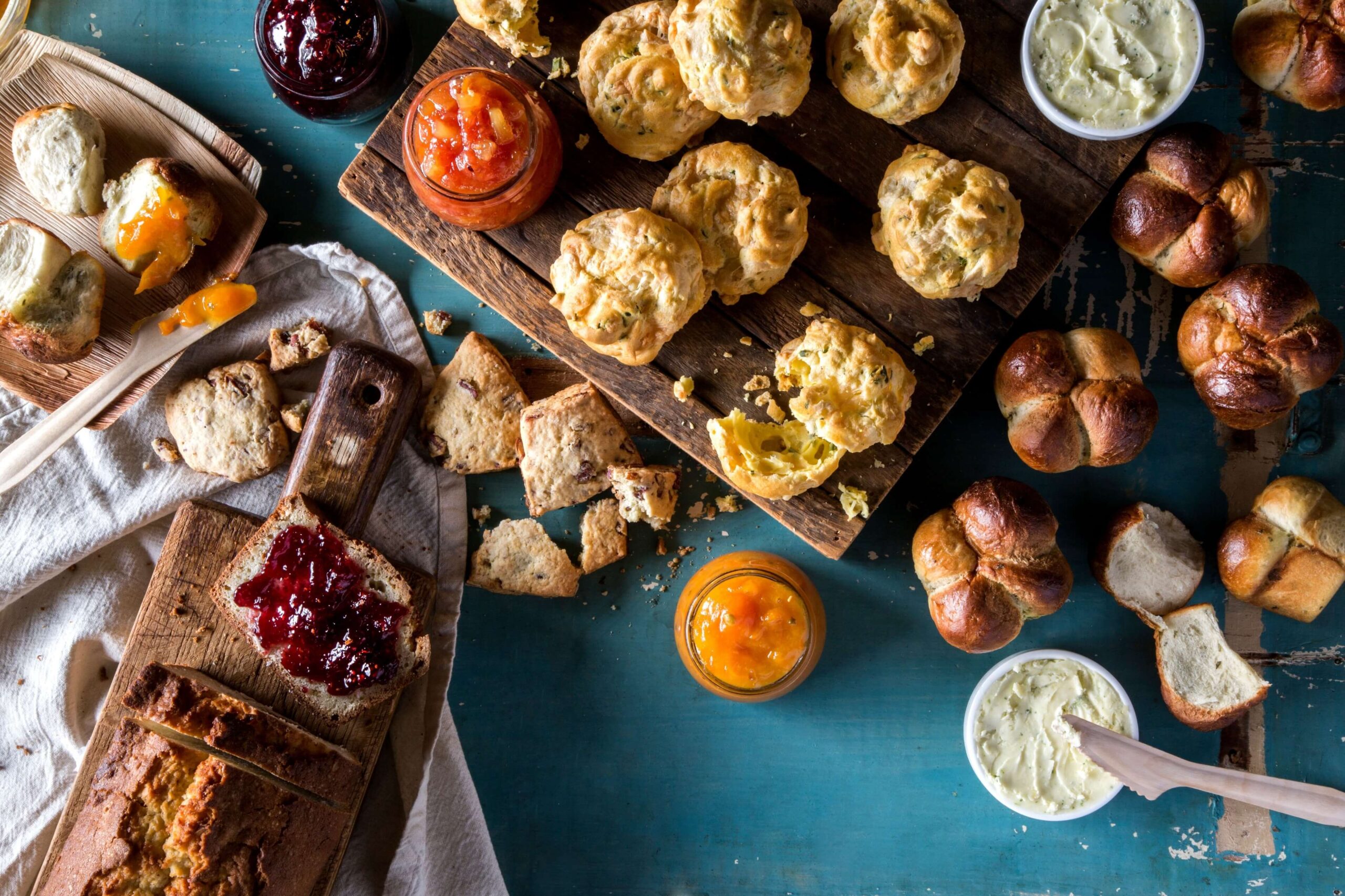 Assorted breads, scones, and muffins are arranged on a table with jars of jam, butter, and cream cheese, plus slices of bread spread with jam.
