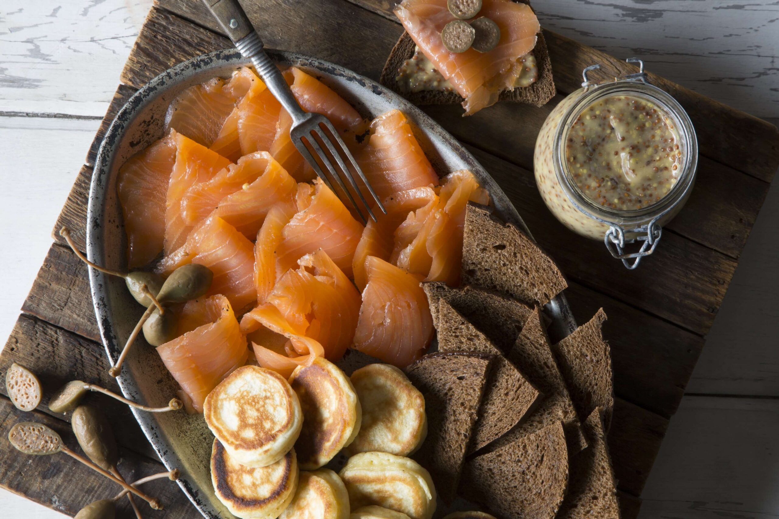 A platter with slices of smoked salmon, dark bread, blini pancakes, caperberries, and a jar of whole grain mustard on a wooden board.