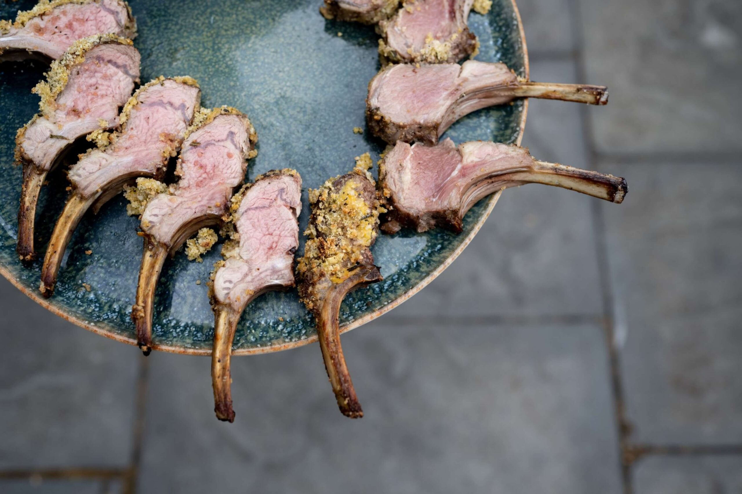 A green plate holds several cooked lamb chops arranged in a semicircle, with pink centers and a breadcrumb crust, on a stone surface background.