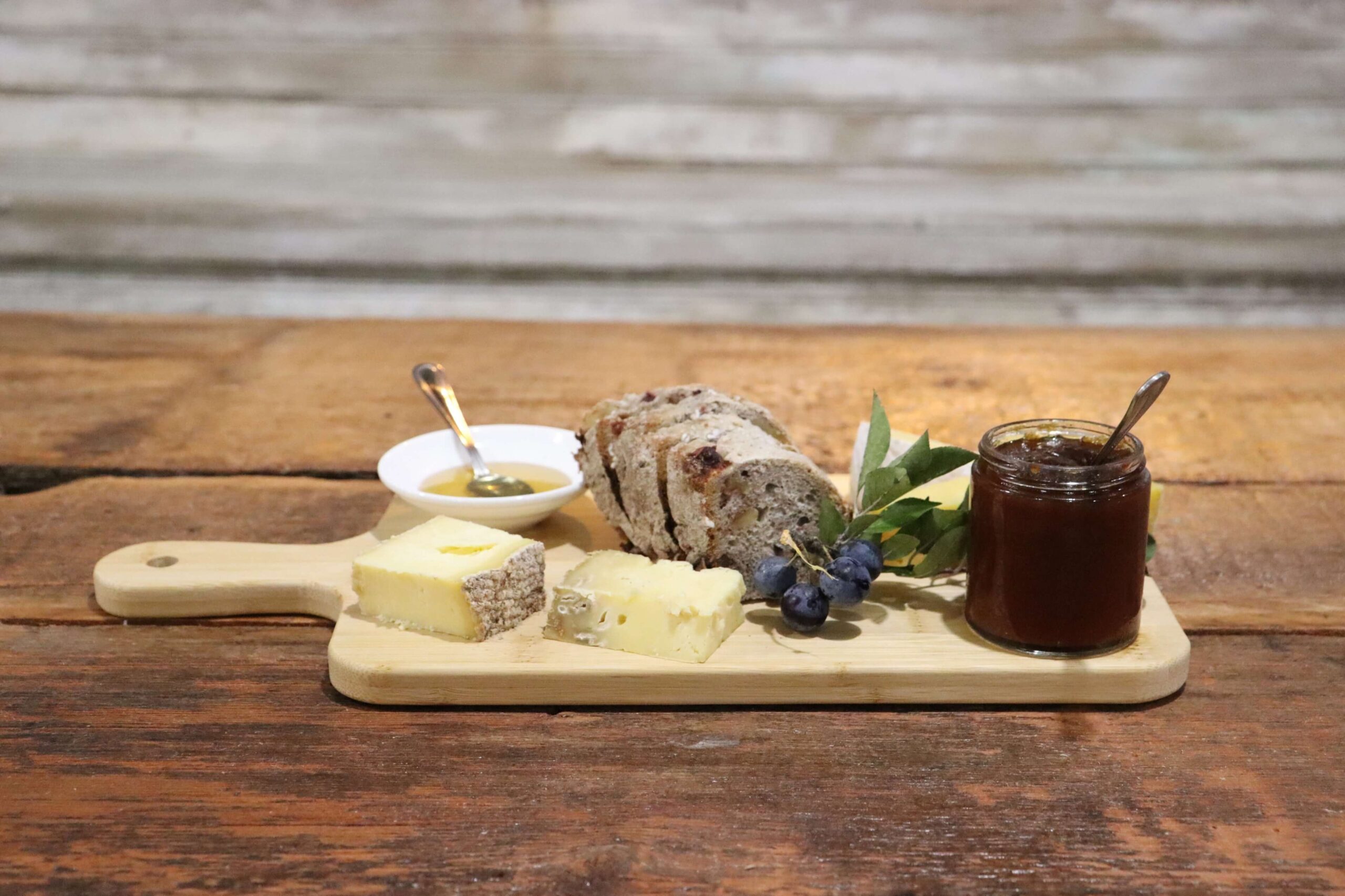 A wooden board with sliced bread, cheese, grapes, a small dish of honey with a spoon, and a jar of preserves on a rustic wooden table.
