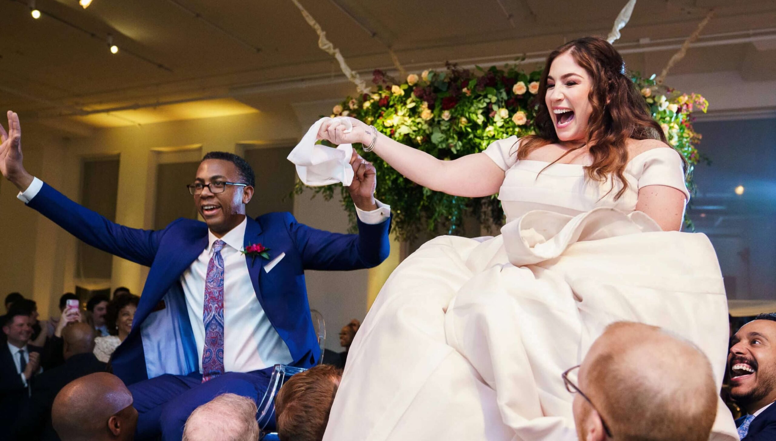 A bride and groom are lifted on chairs during a wedding celebration, smiling and holding a napkin, surrounded by guests indoors.