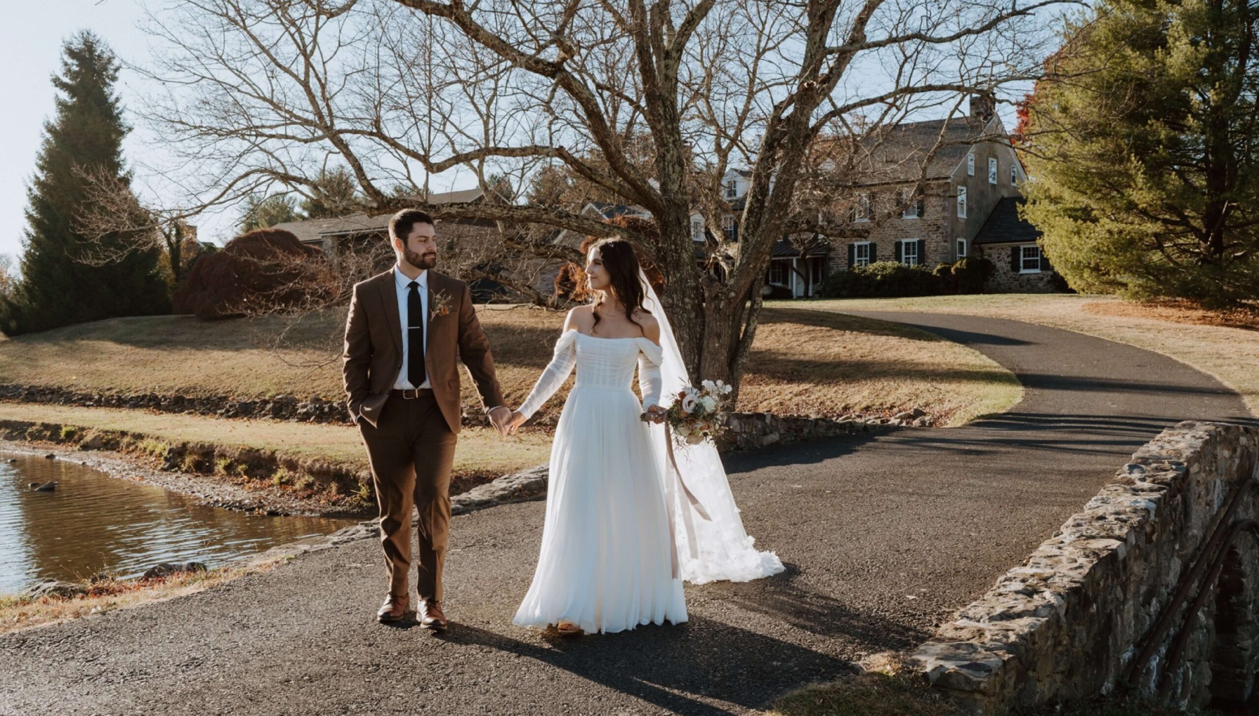 A bride and groom walk hand in hand on a paved path near a pond, with trees and a large house in the background on a sunny day.