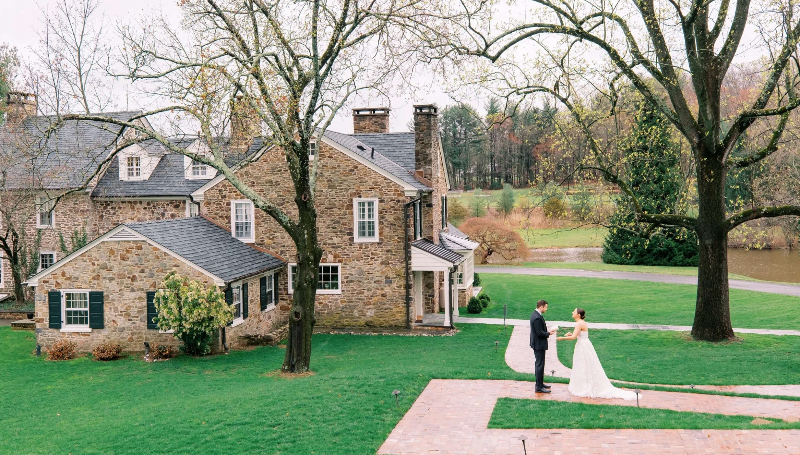 A bride and groom stand holding hands on a brick path outside a large stone house with green lawns and leafless trees.
