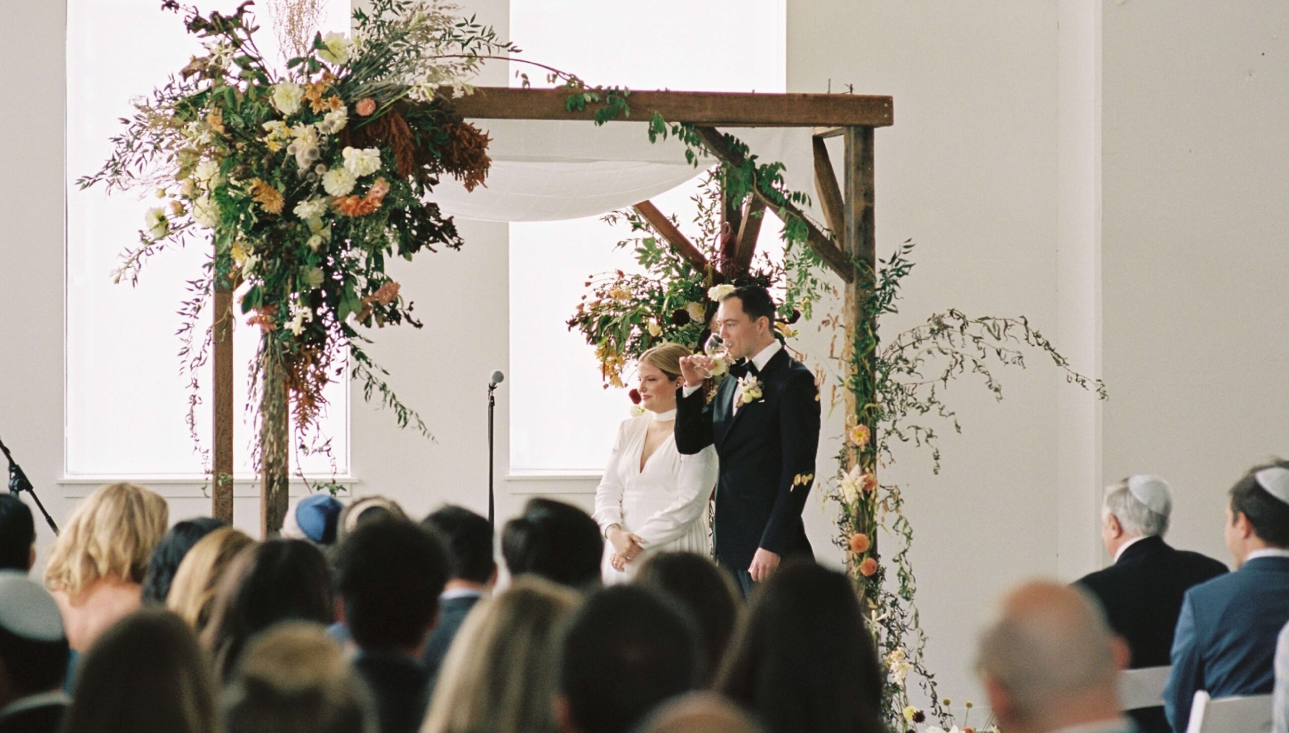 A bride and groom stand under a floral chuppah during a wedding ceremony, facing a seated audience while the groom drinks from a cup.
