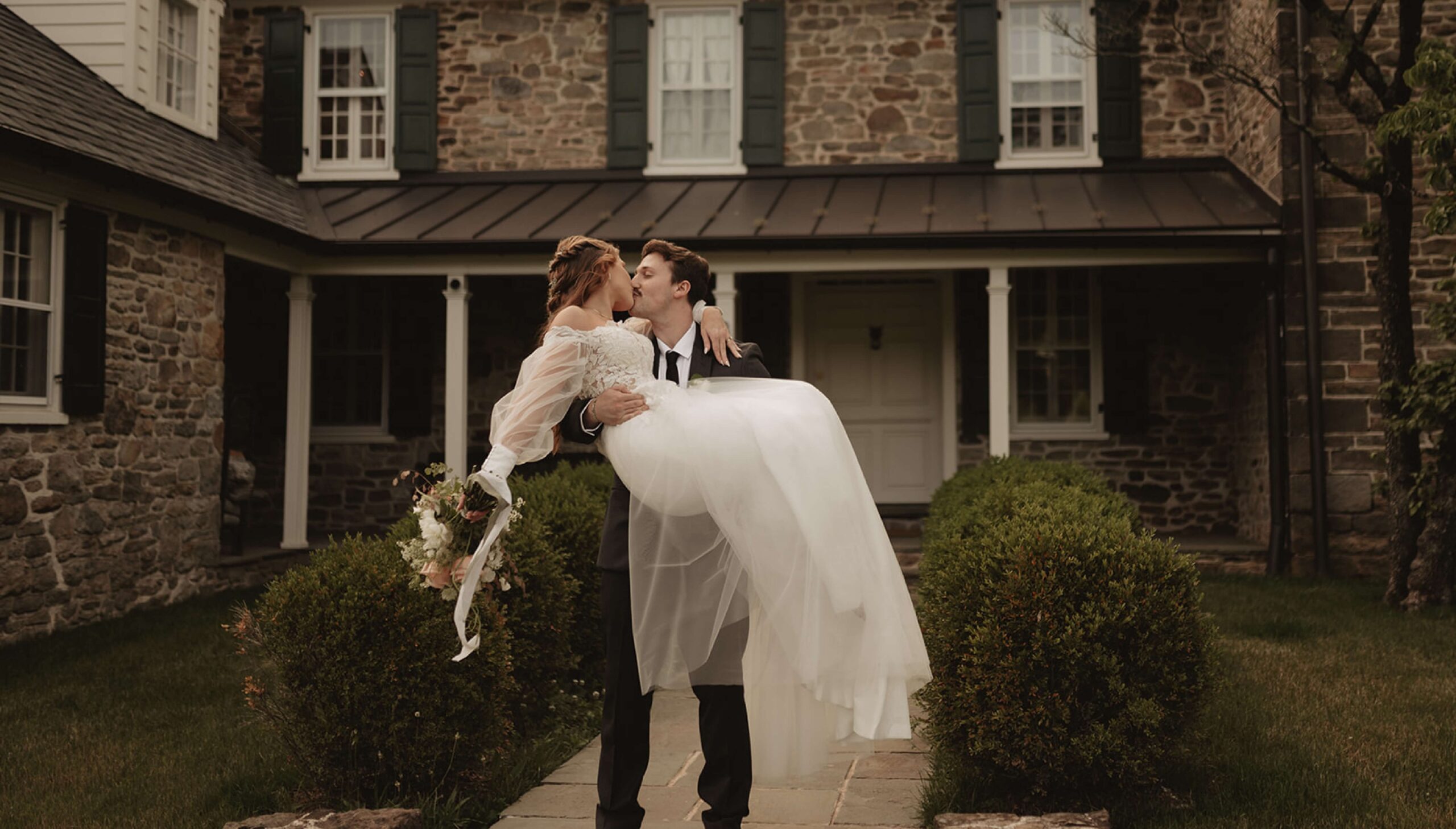 A groom in a suit carries a bride in a white dress and holding flowers outside a stone house with green shutters.