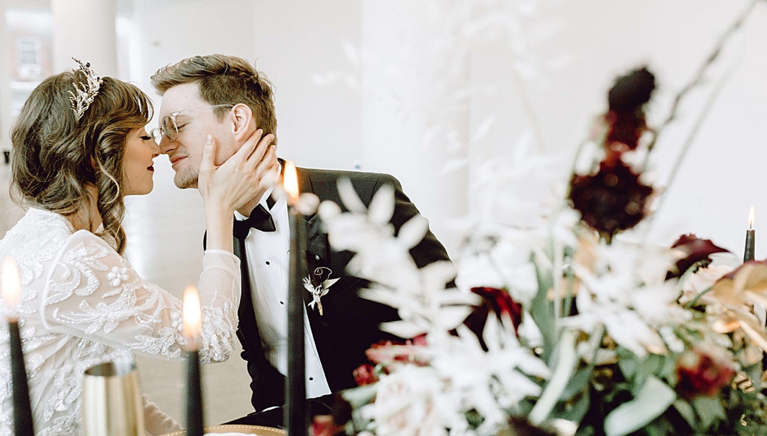 A bride and groom sit closely, about to kiss, at a decorated table with candles and floral arrangements in a bright, modern venue.