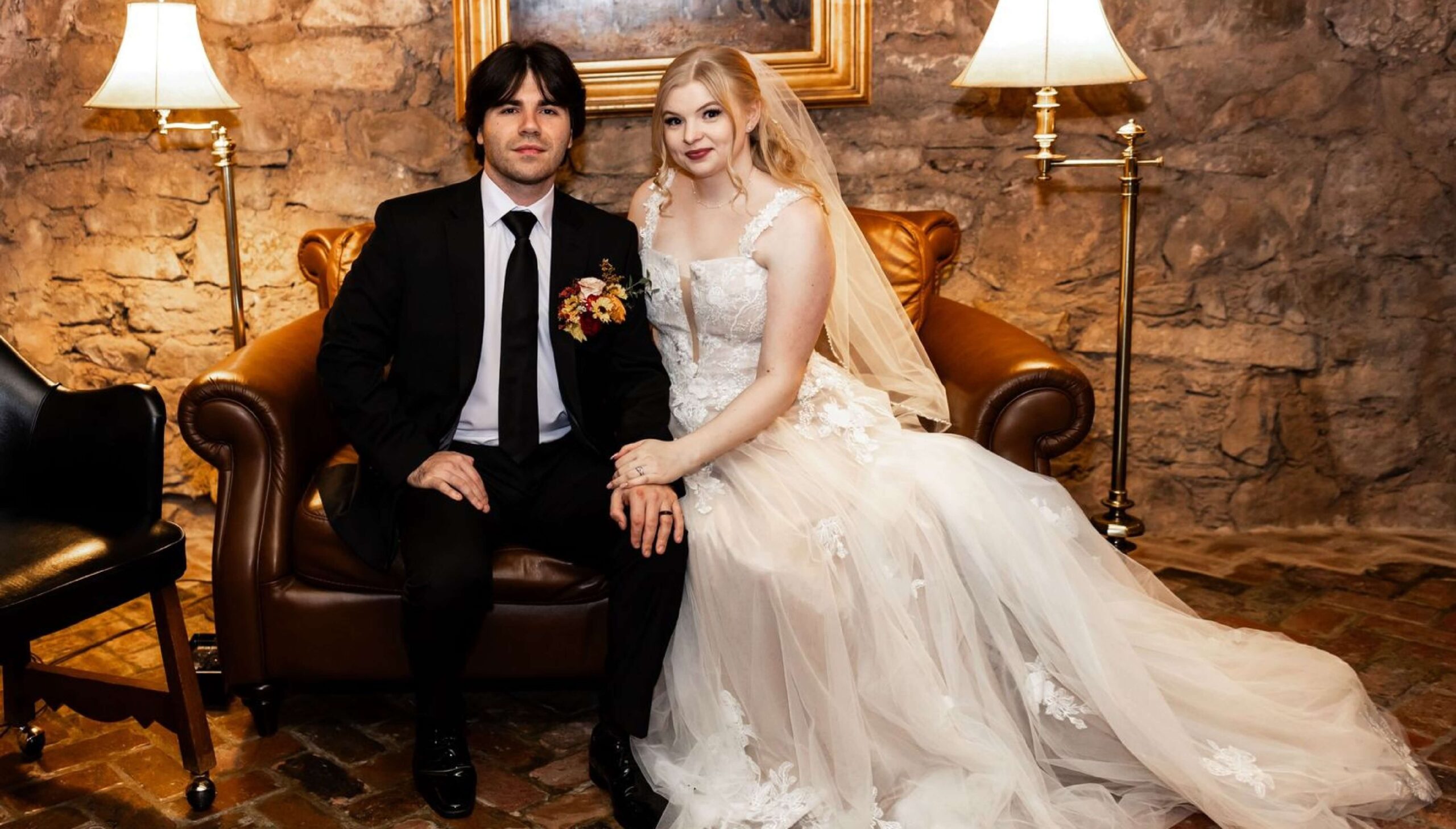 A bride in a white wedding dress and a groom in a black suit sit together on a brown leather chair in a rustic room with stone walls and lamps.