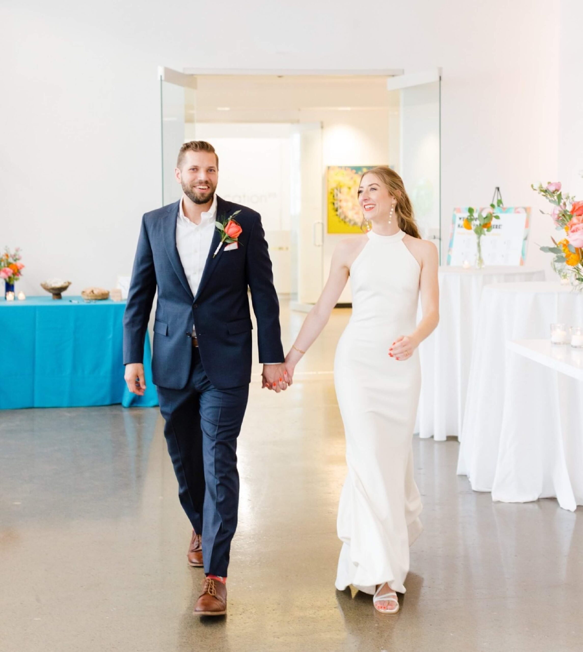 A man in a navy suit and a woman in a white sleeveless dress walk hand in hand indoors, possibly at a formal event or wedding reception.