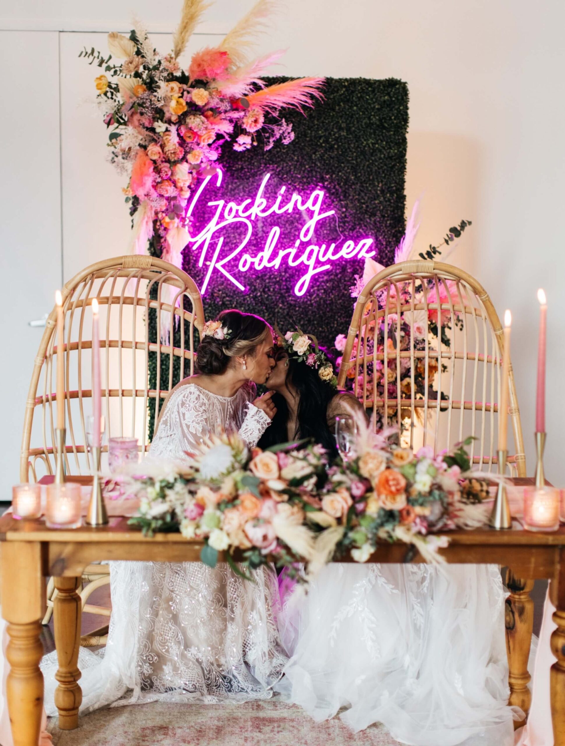 Two brides in white dresses share a kiss at a decorated wedding table with flowers and candles, beneath a neon “Gocking Rodriguez” sign and floral backdrop.